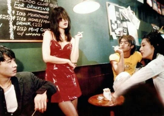 A woman in a red dress standing inside a cafe or restaurant, with three seated men nearby, two of whom are smoking.