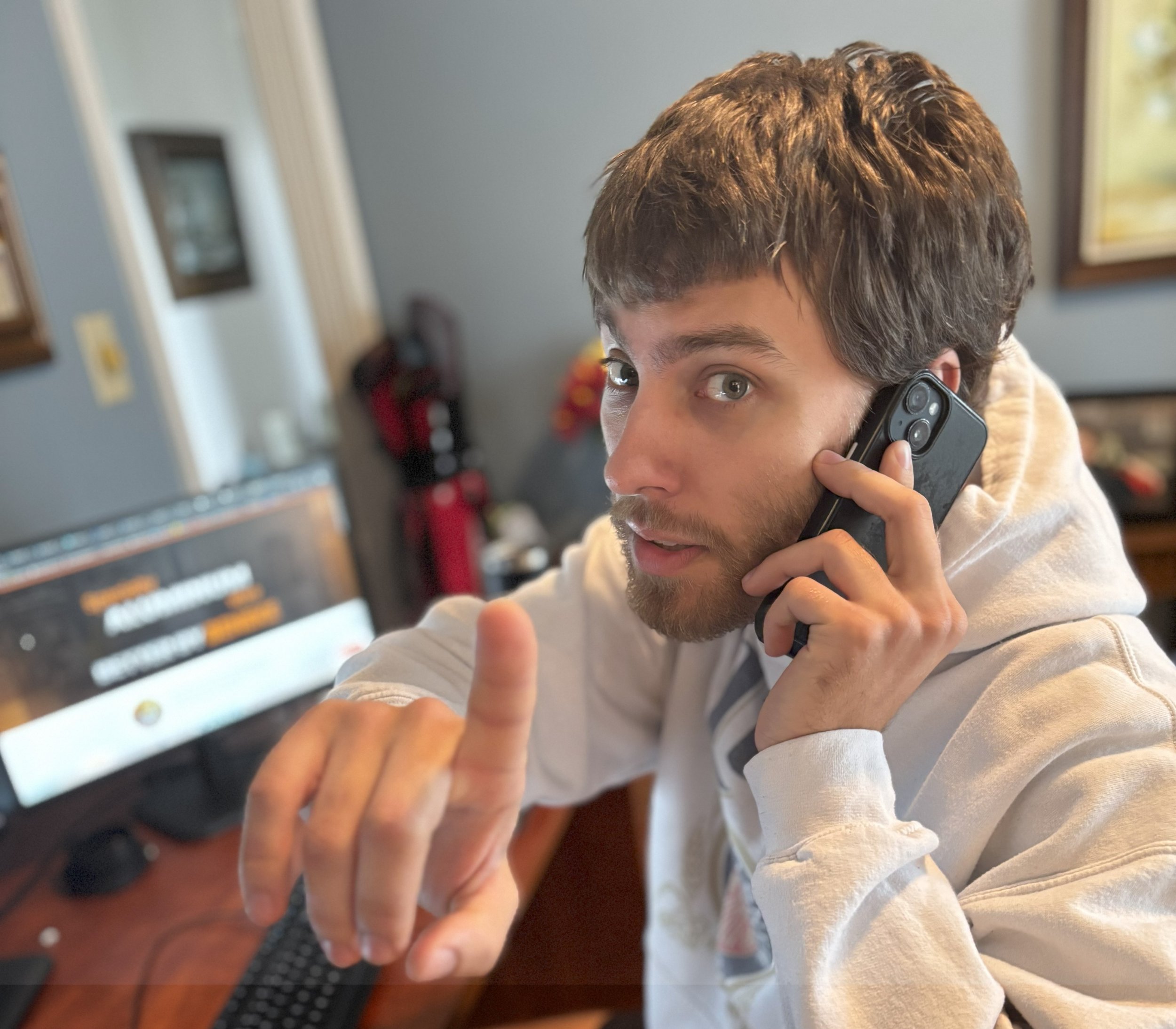 A young man with brown hair and a beard holding a phone to his ear, pointing his finger upward, sitting in a home office or living room.