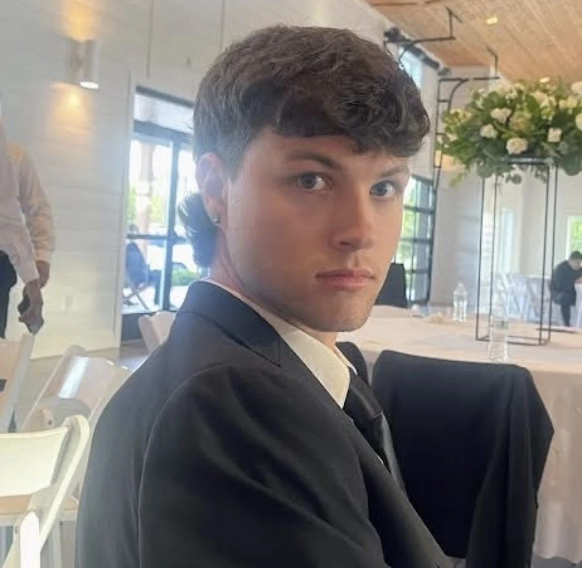 Young man with short dark hair wearing a black suit, white shirt, and black tie, sitting in a well-lit indoor setting with flowers and people in the background.