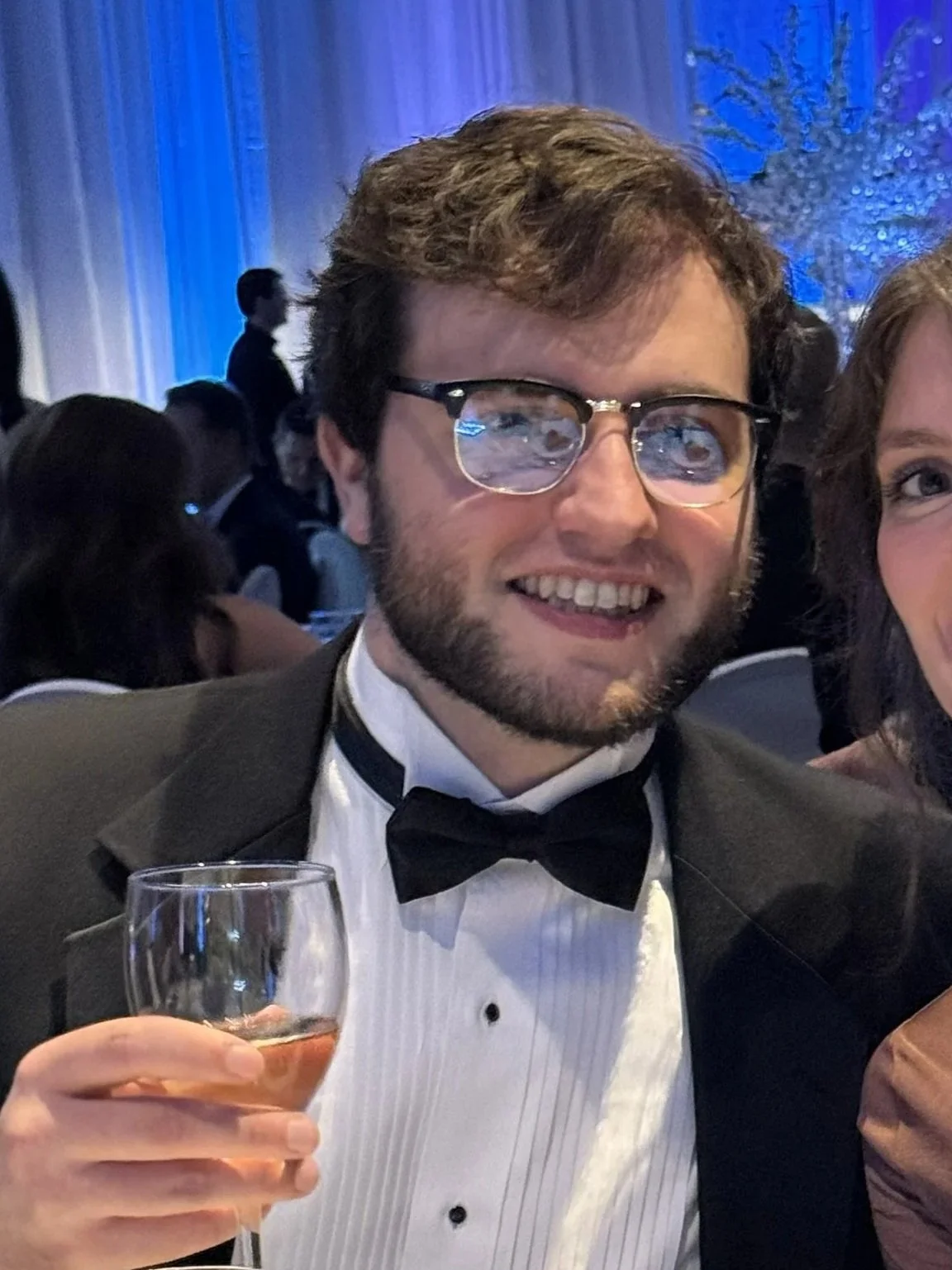 A man in formal tuxedo with a bow tie and glasses holding a wine glass at a formal event or celebration.