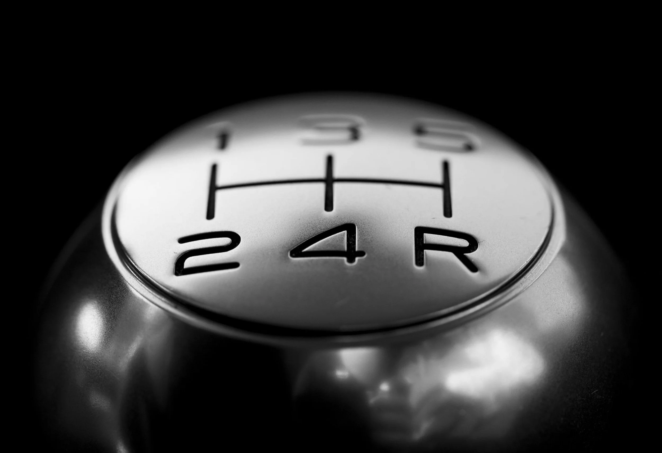 Close-up of a manual car gear shift knob with the gear pattern and numbers visible, in black and white.