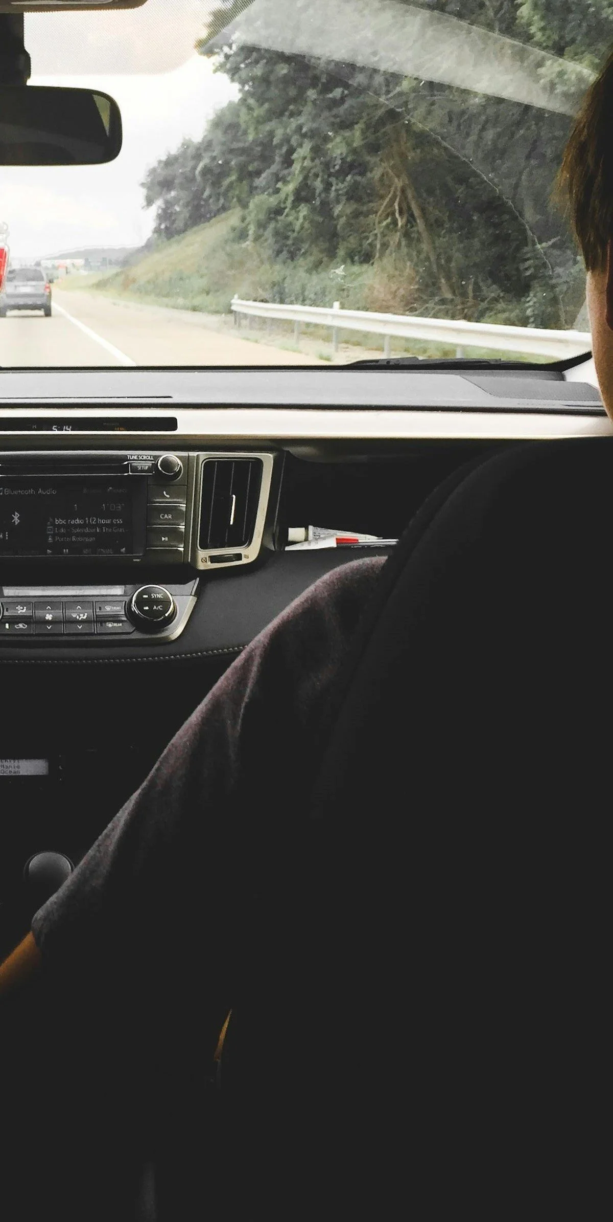 Inside a moving car, view from the passenger seat shows the dashboard with a car radio, climate control, and side mirror. Through the windshield is a highway with a car ahead and a tree-lined hillside on the side.