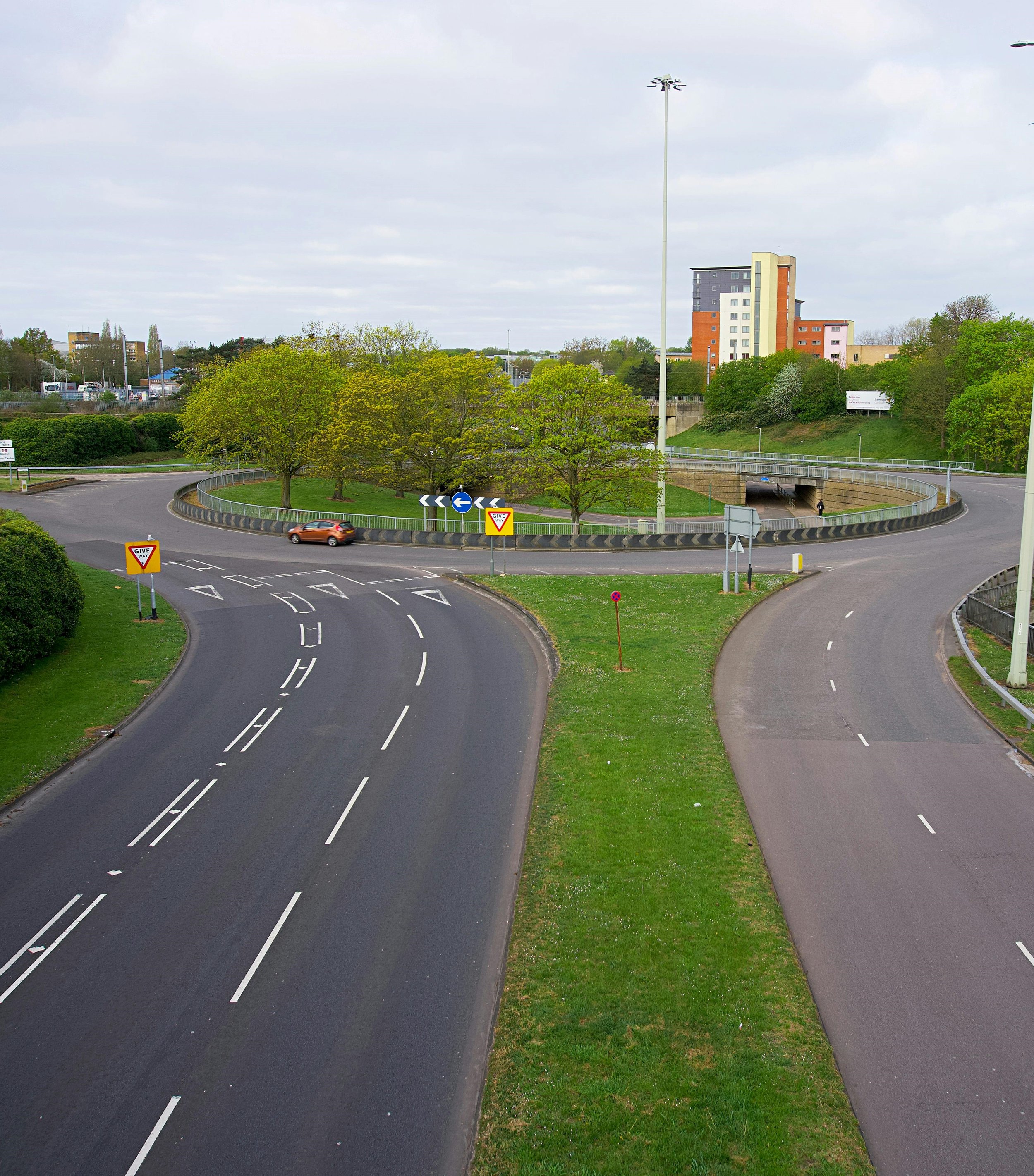A circular roadway with multiple lanes separated by white dashed lines and a grassy median, featuring traffic signs and a car. Surrounding greenery and trees, with a cityscape in the background.