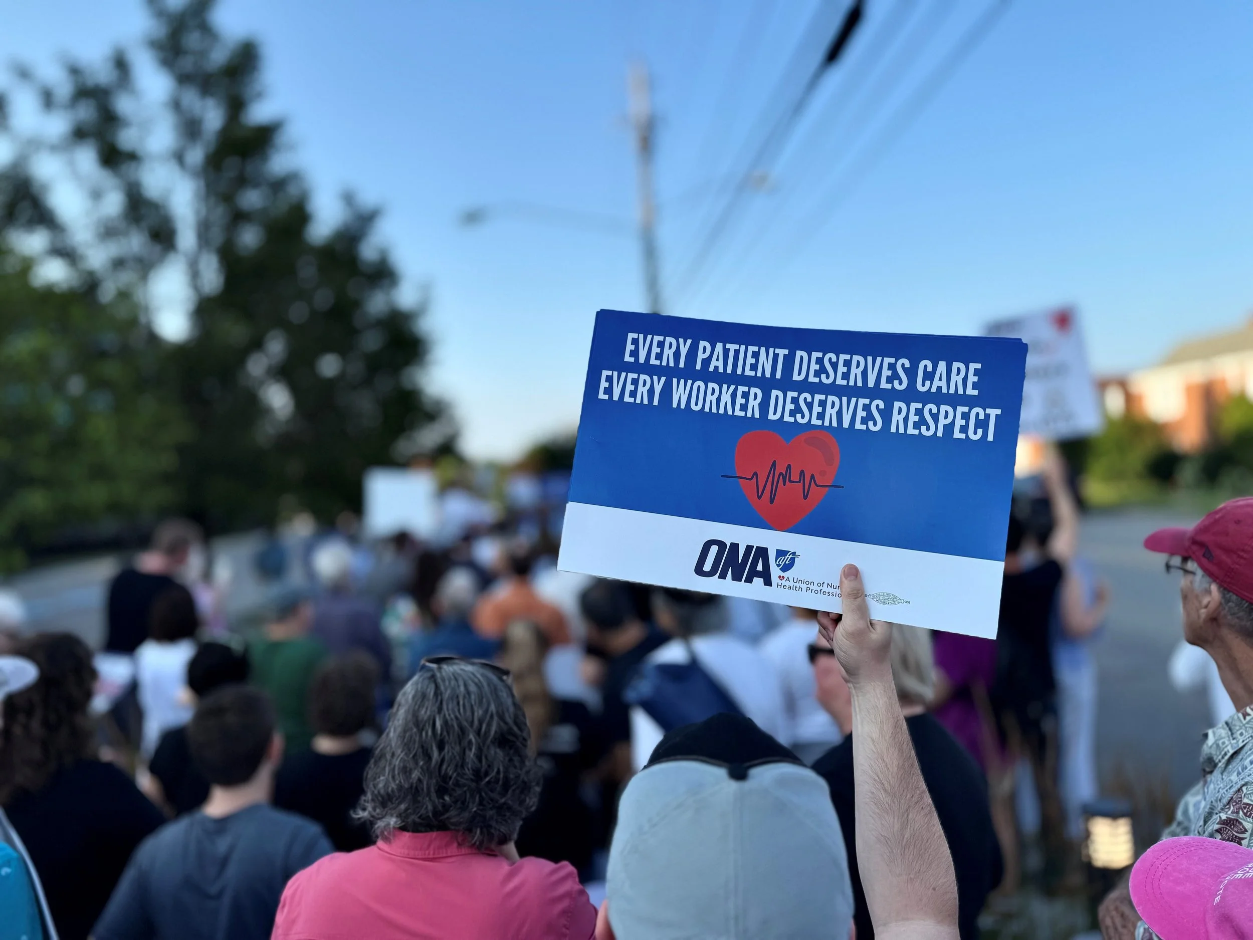 A protest or rally with a crowd of people holding signs, one sign visible in the foreground reads, 'Every patient deserves care, every worker deserves respect,' organized by the ONA union.