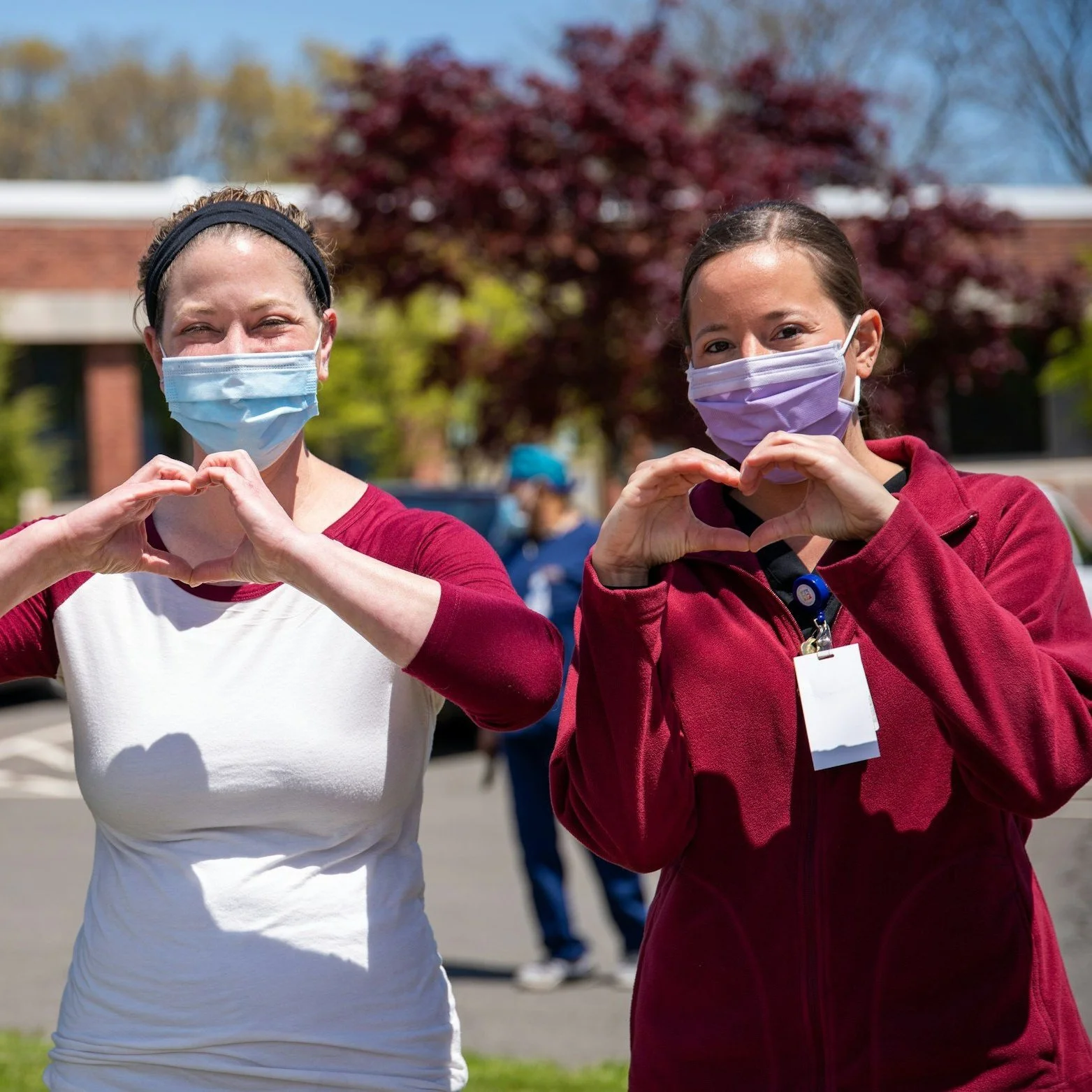 Two women wearing face masks making heart signs with their hands outdoors on a sunny day.