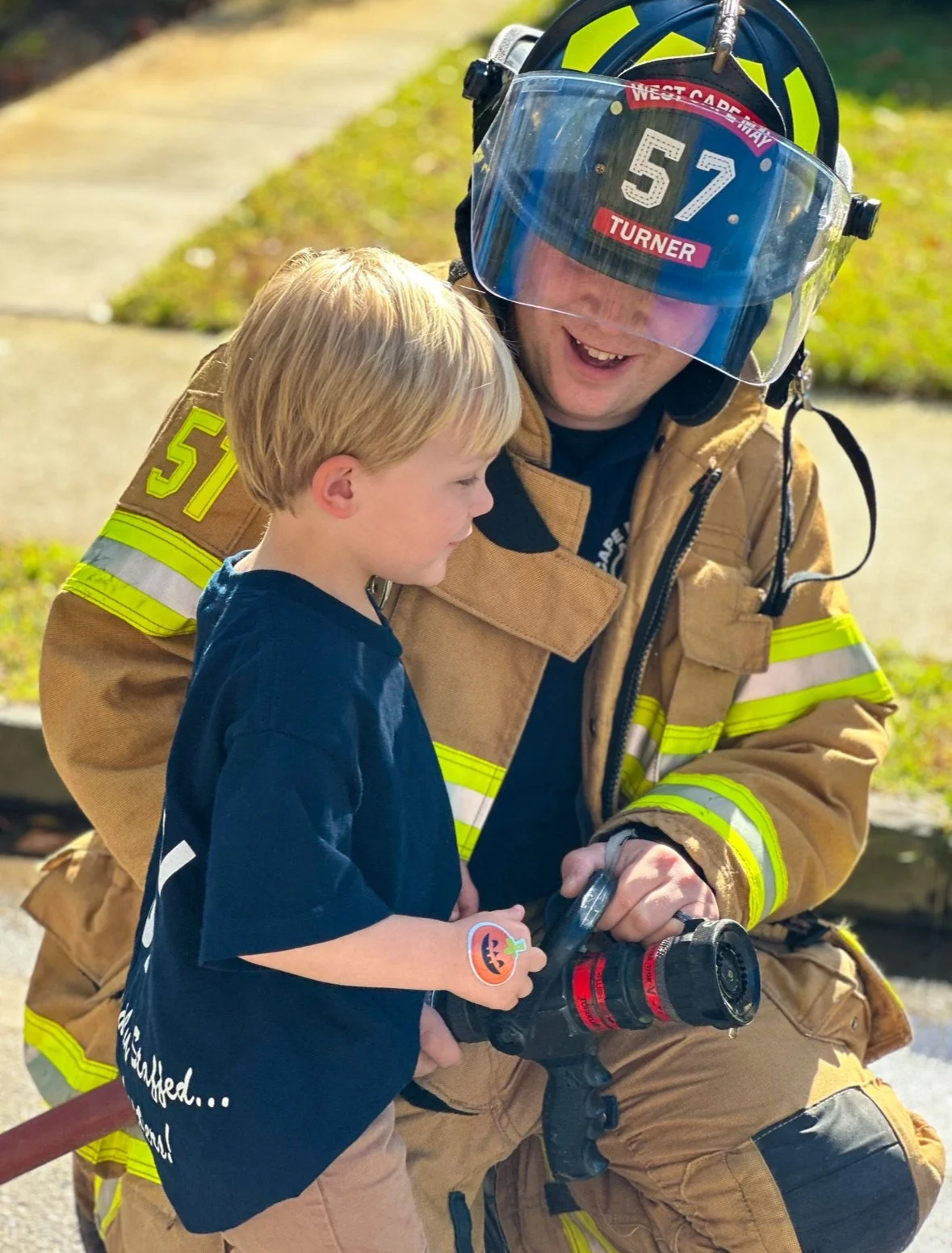 A firefighter in full gear, including a helmet with the number 57 and a face shield labeled 'West Cabr' and 'Turner,' is sitting outdoors with a young boy. They are both focused on a fire hose nozzle the firefighter is holding, and the boy has a pump