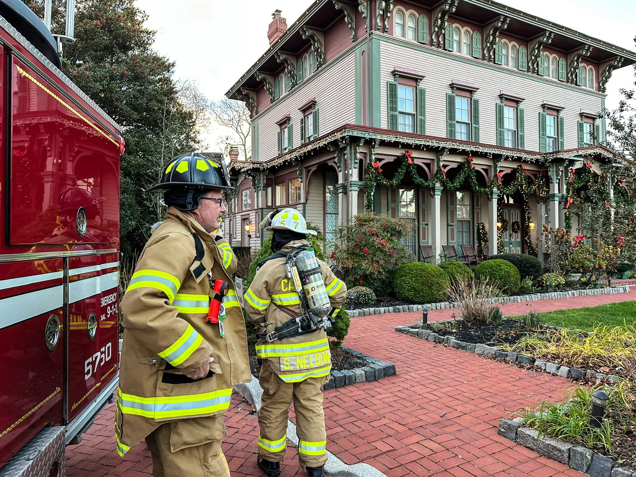 Two firefighters in full gear standing outside a large, Victorian-style house decorated with holiday lights and wreaths. A red fire truck is parked nearby.