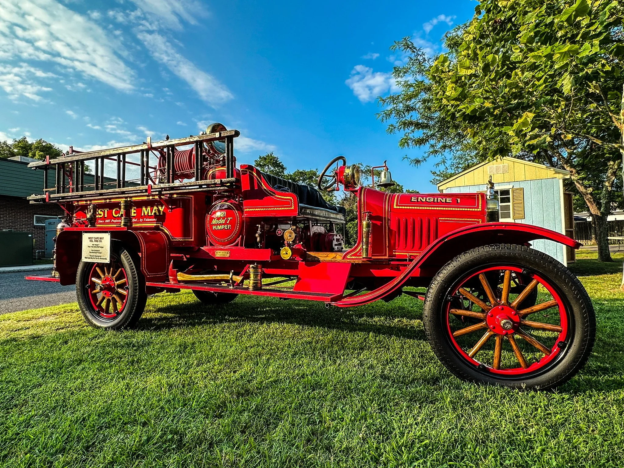 Vintage red fire truck with wooden wheels parked on green grass next to a small house and trees under a blue sky with scattered clouds.