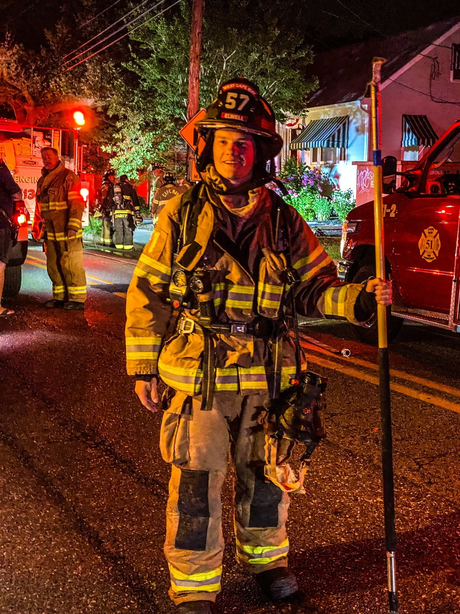 Firefighter standing on wet street at night, holding a yellow tool, with other firefighters and emergency vehicles in the background.