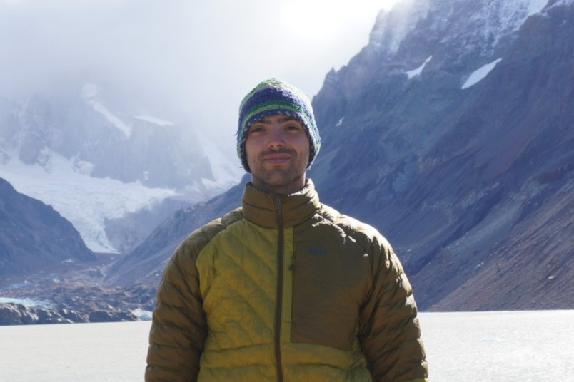 A man wearing a winter hat and a brown jacket standing in front of a snowy mountain landscape with glaciers.