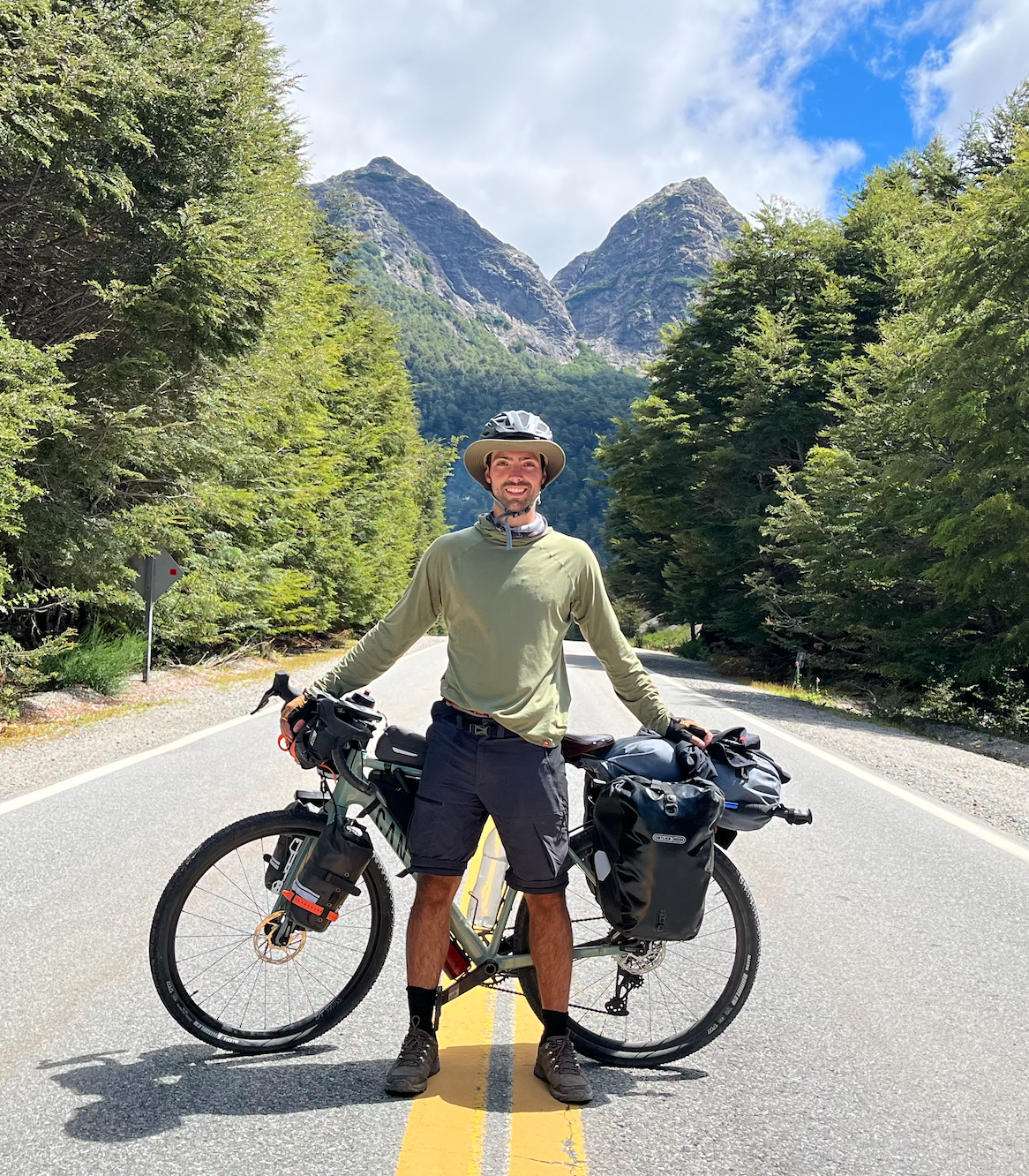 Man standing with a loaded touring bicycle on a forested mountain road, smiling, wearing a helmet and outdoor clothing.