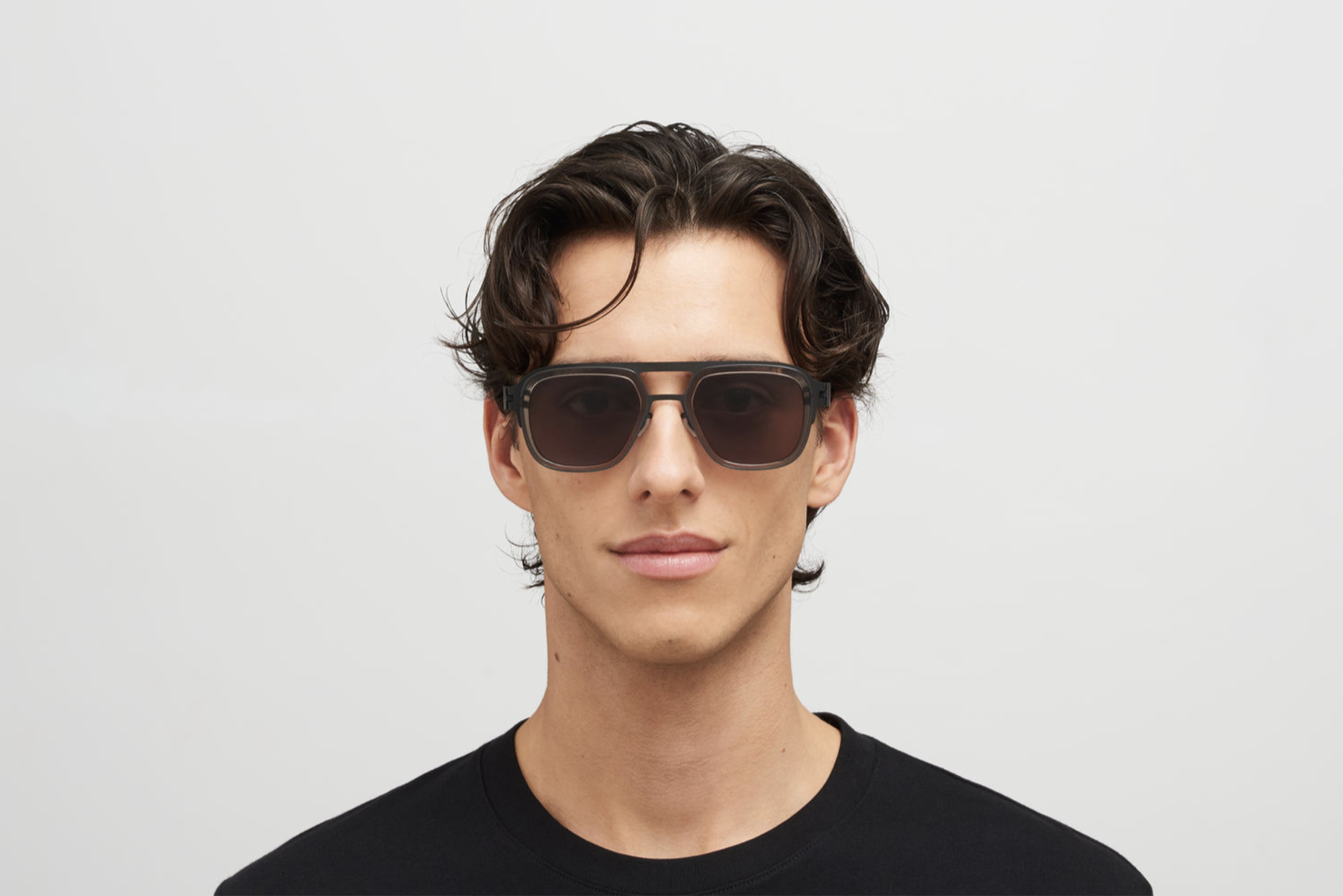 Young man with wavy brown hair wearing black sunglasses and a black shirt, facing forward against a plain light gray background.