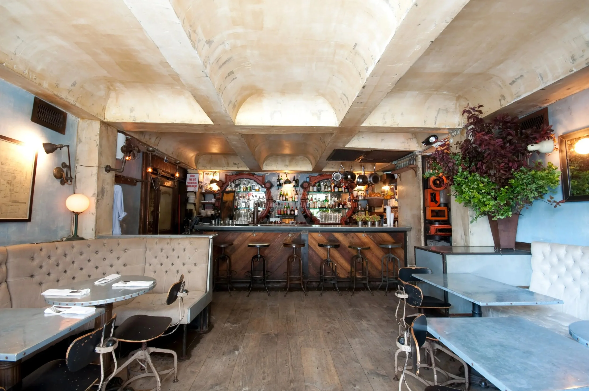 Interior view of a cozy bar with a marble top counter, bar stools, and a well-stocked back bar with bottles and glasses. There are tables and chairs in the dining area, with a large potted plant and framed artwork on the walls. The ceiling has a text