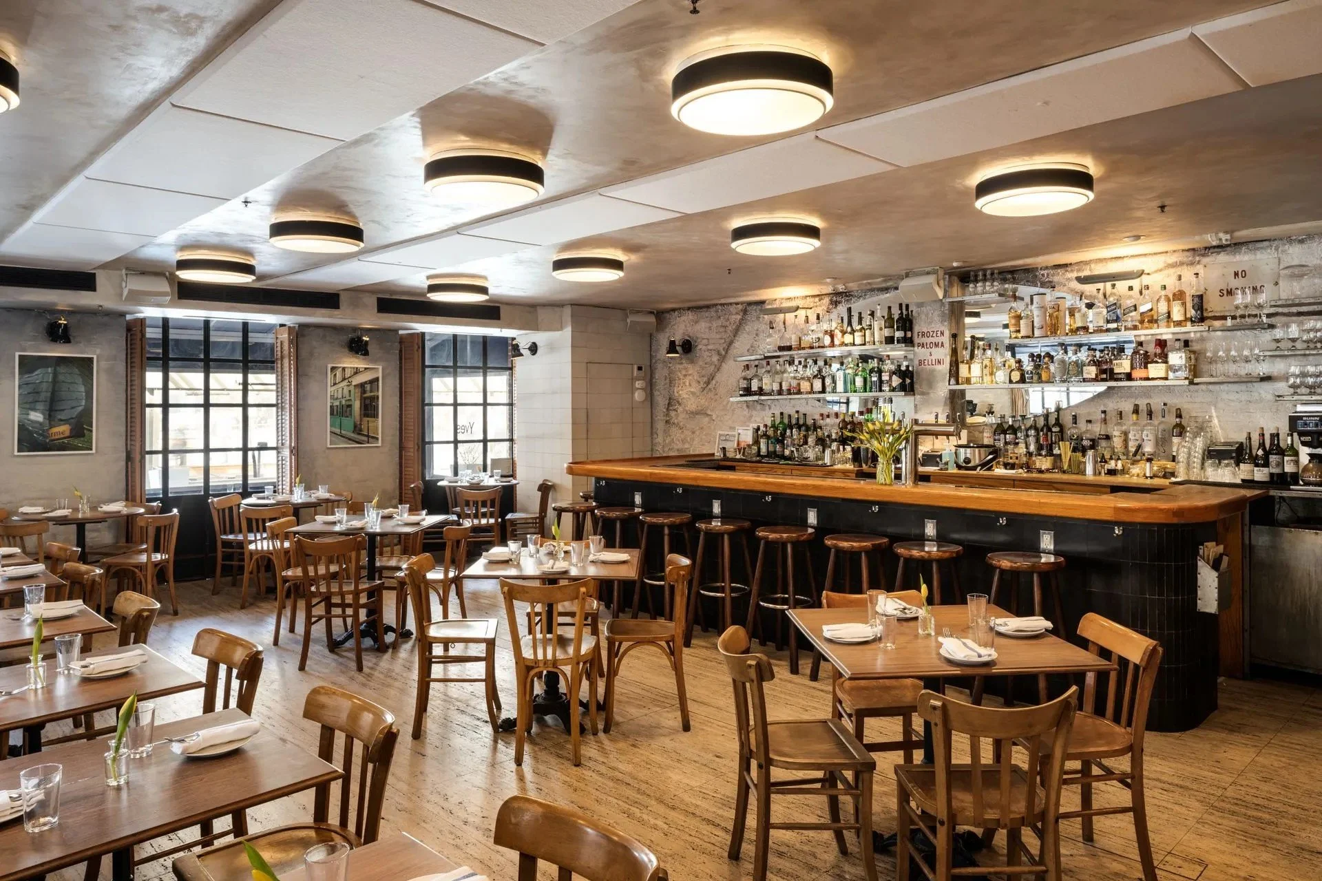 Upscale restaurant with wooden tables and chairs, a bar with alcohol bottles, hanging ceiling lights, large windows, and minimal decor.