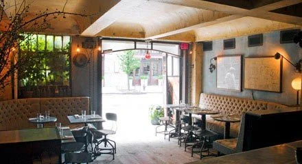 Interior of a cozy restaurant with booths, tables, and chairs near the entrance, warm lighting, and a view outside through open doors.