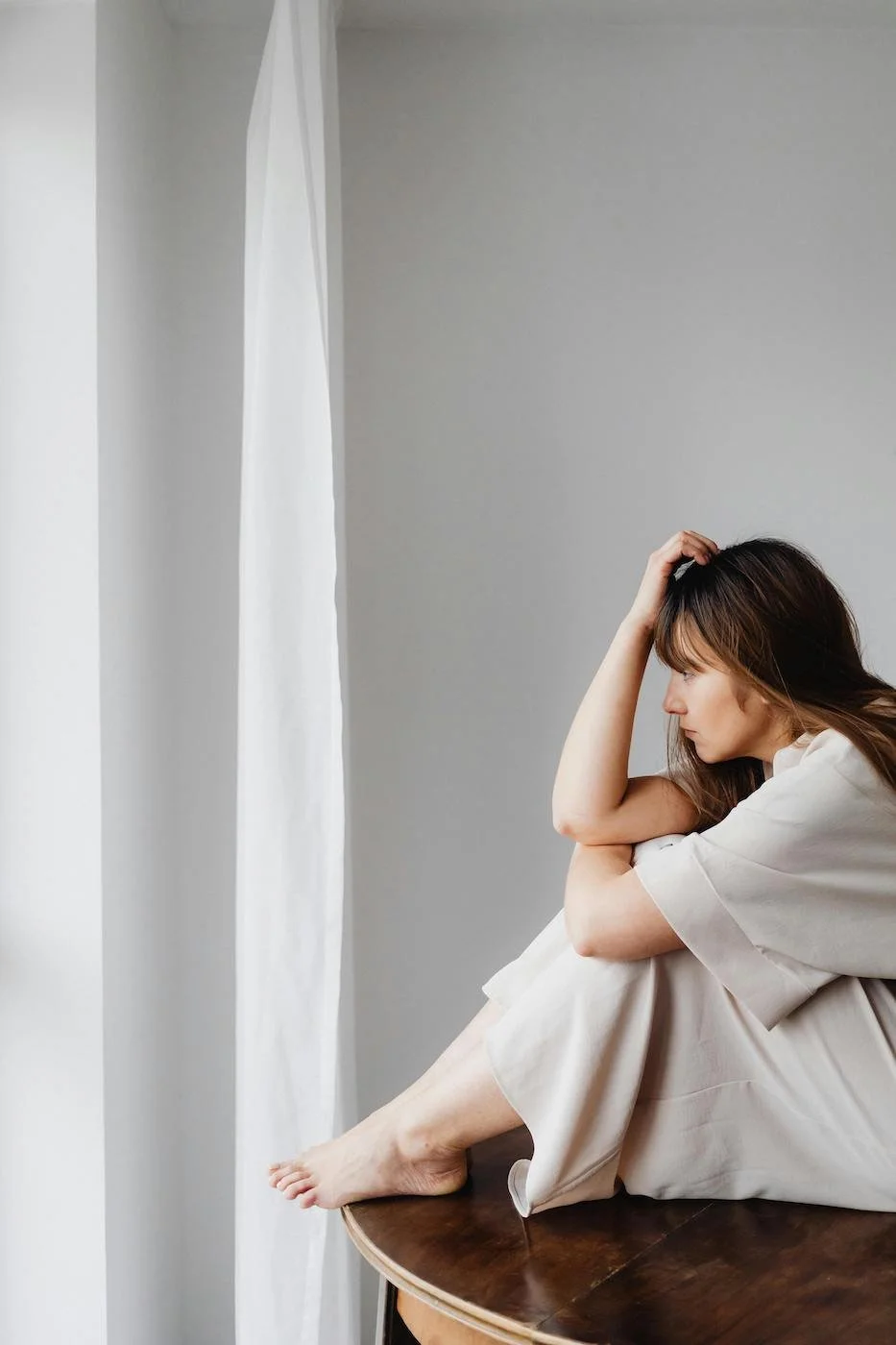 A woman sitting on a wooden table, looking down with a thoughtful or sad expression, with her head resting in her hand, wearing light-colored pajamas, near a white curtain - to represent how patients of Nicole Wrightson Naturopath may be feeling