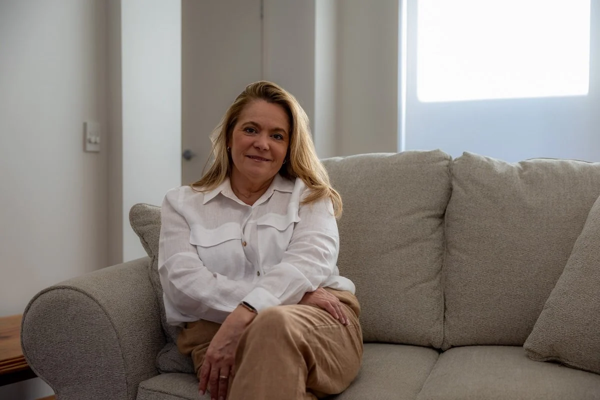 Naturopath Nicole Wrightson from Natured Wellness sits on a beige couch in a well-lit living room, wearing a white blouse and tan pants.