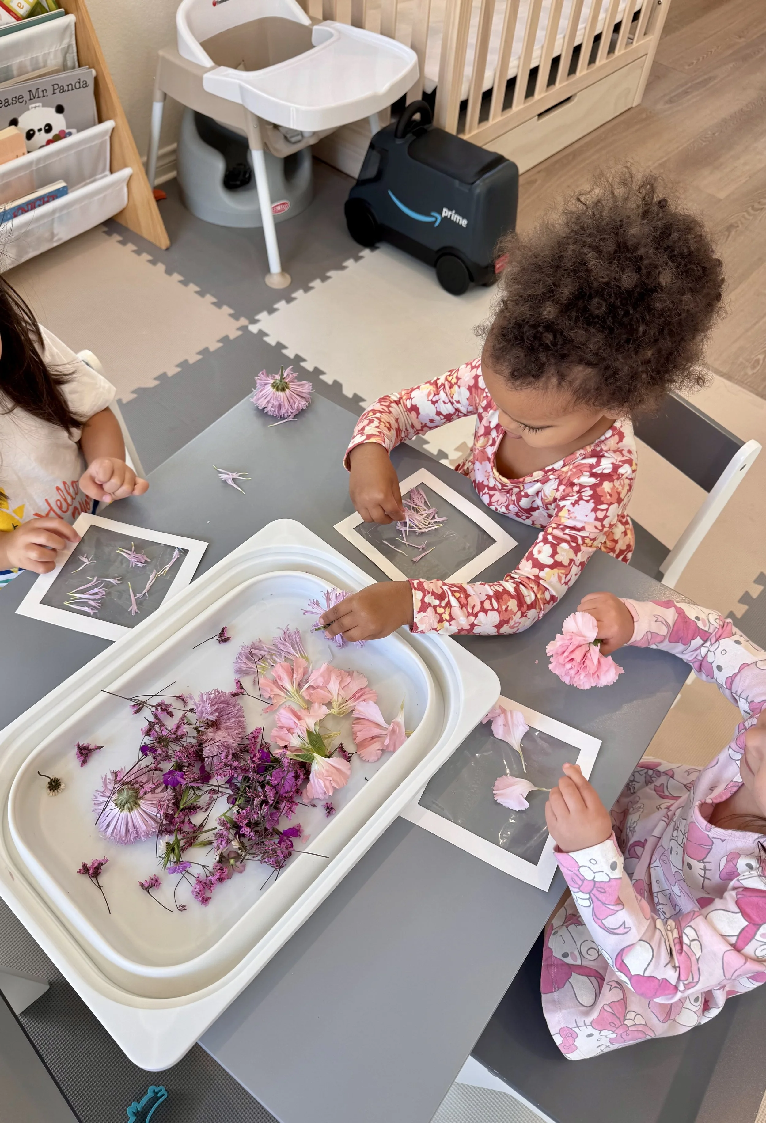 Two children sitting at a table with pictures and flowers, arranging pink and purple flowers on paper, with a collection of flowers in a white tray in front of them.