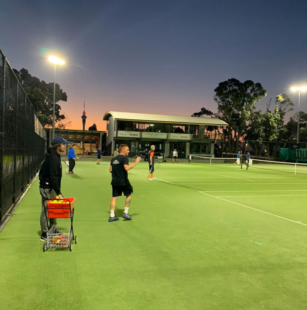 People playing tennis on an outdoor court during sunset, with some individuals preparing and others actively playing, surrounded by trees and outdoor lighting.