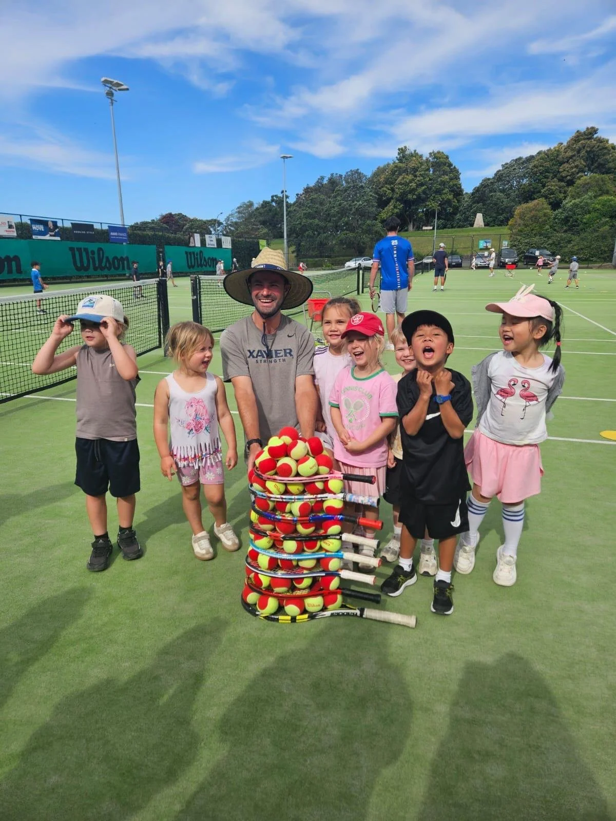 Group of children and a coach on a tennis court with tennis balls and rackets, smiling and celebrating.