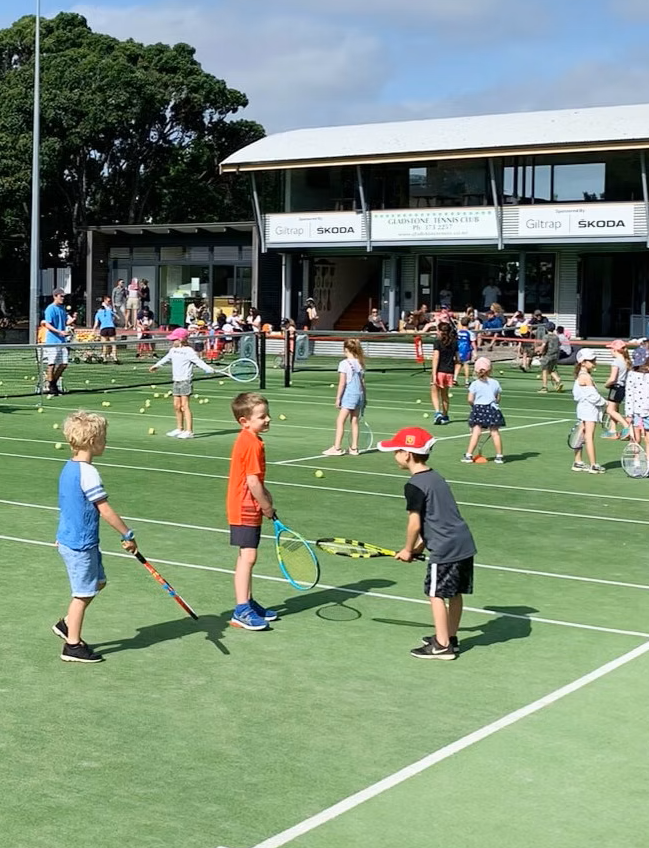 Children playing tennis and practicing with rackets on a tennis court during daytime, with a large clubhouse and trees in the background.