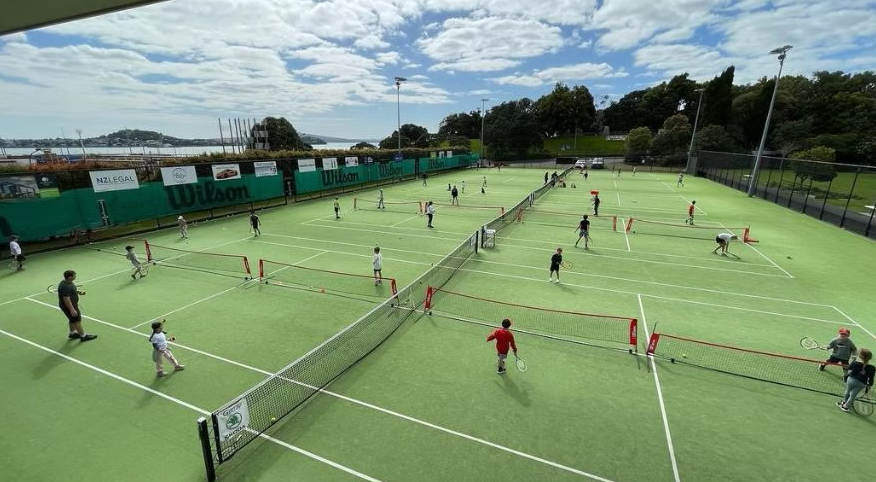 People of various ages practicing tennis on multiple courts with green surfaces and surrounded by a black fence, under a partly cloudy sky.
