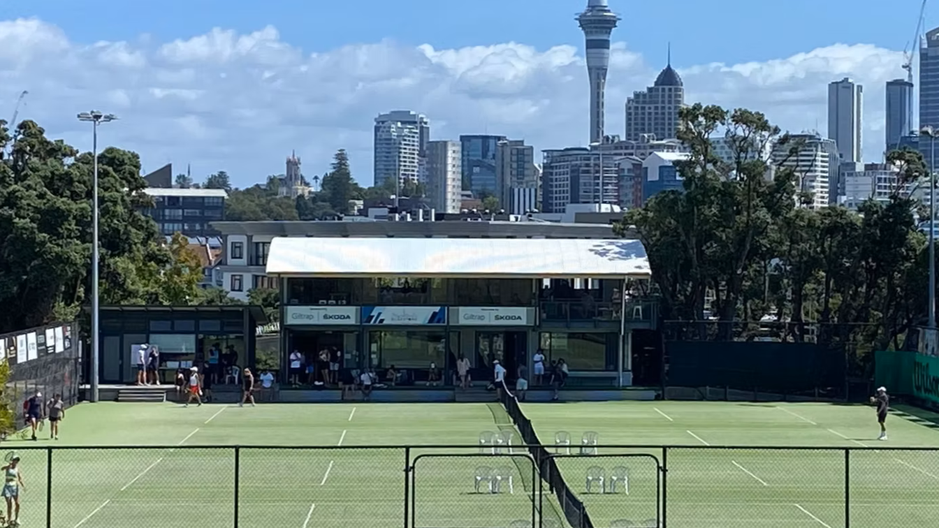 Outdoor tennis courts with players and spectators, city skyline in the background, and a two-story building with sponsors