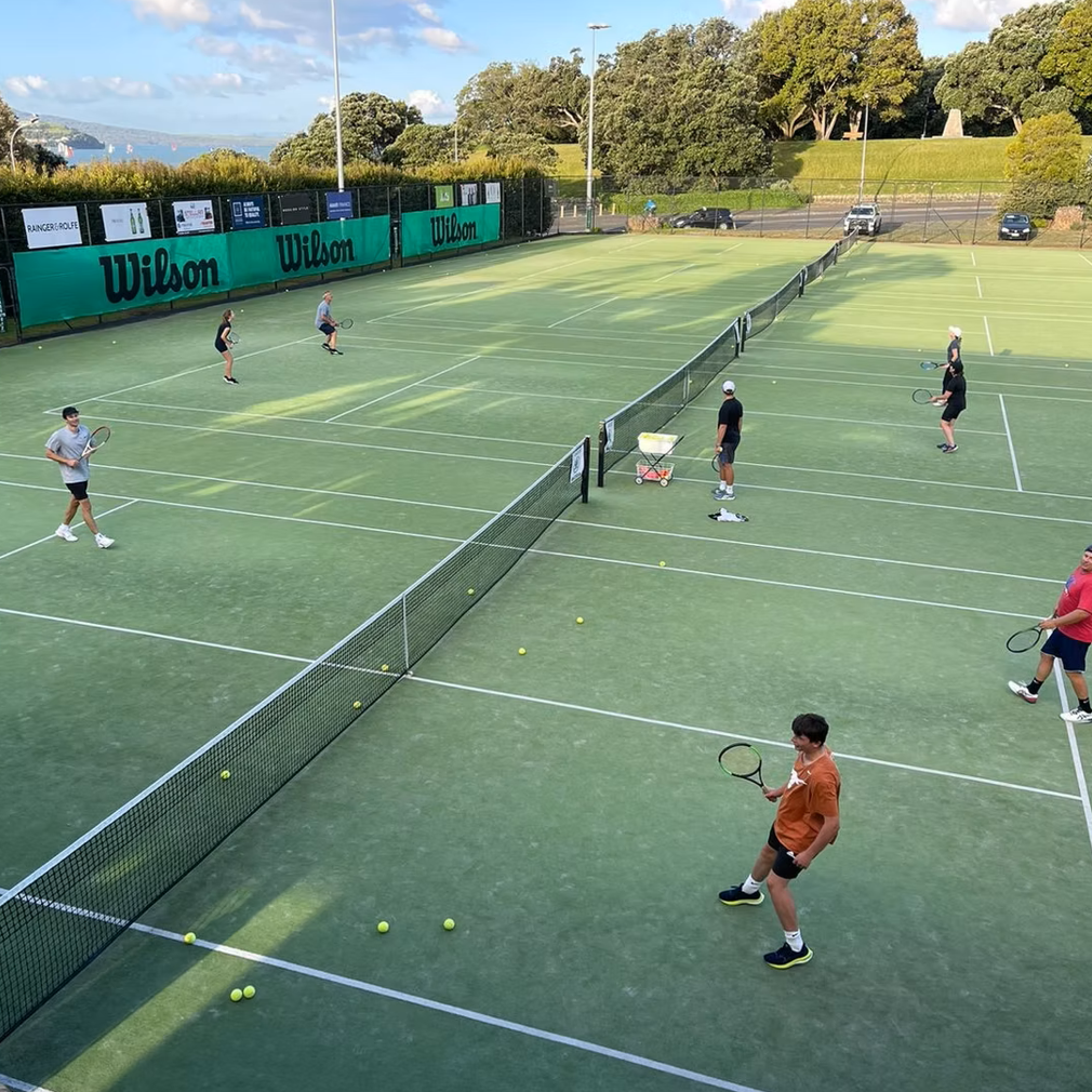 People playing tennis on an outdoor court with a Wilson advertisement backdrop, some in casual sportswear and tennis equipment, during daytime with a partly cloudy sky.
