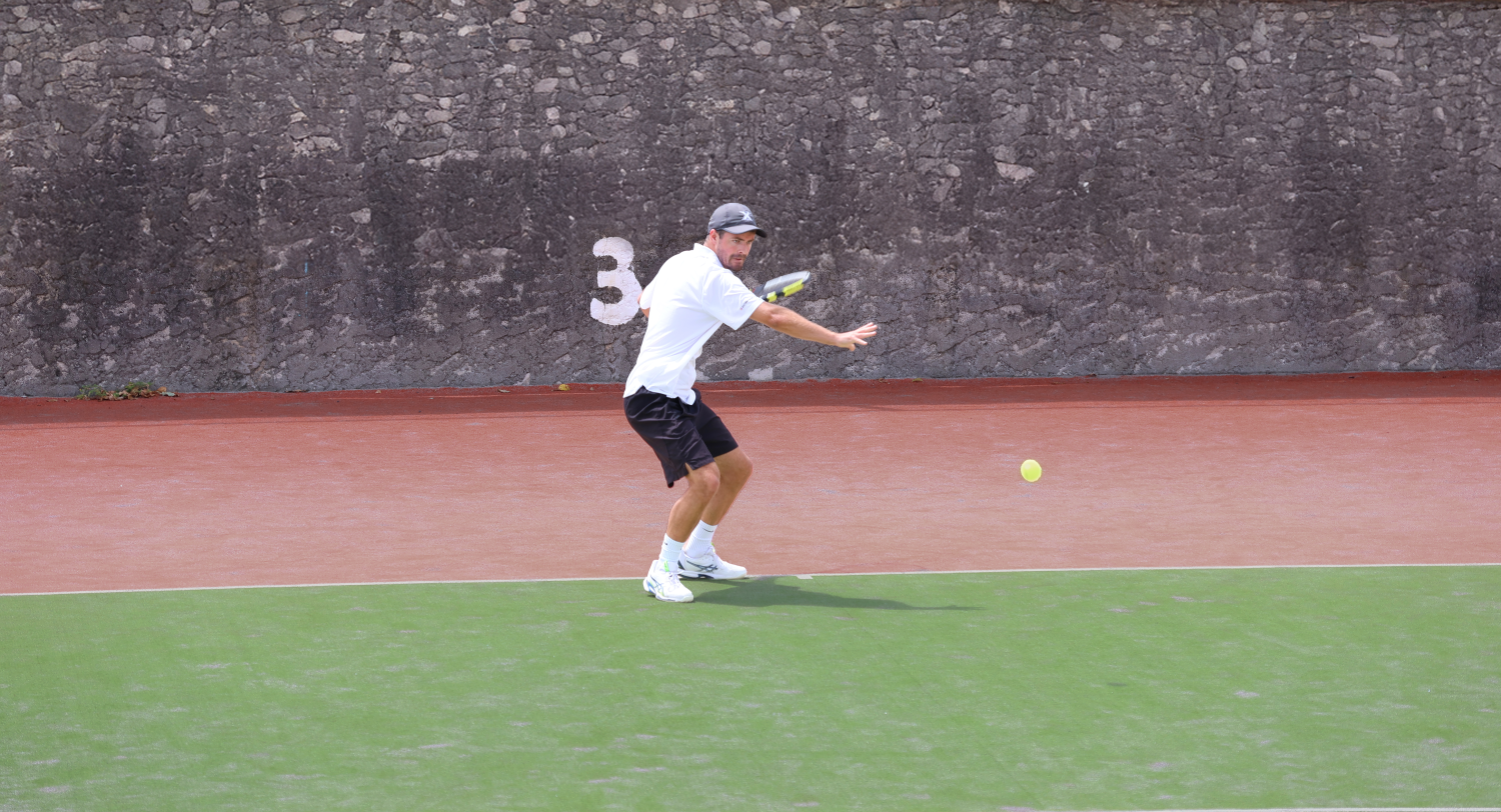 A man playing tennis on a court, preparing to hit a tennis ball with a racket. He is wearing a white shirt, black shorts, white shoes, and a grey cap.