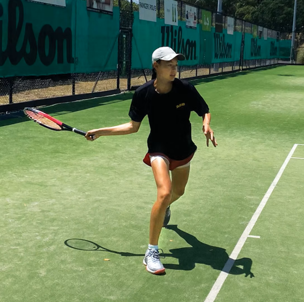 A woman playing tennis on an outdoor court, wearing a white cap, black shirt, red shorts, and gray shoes, holding a tennis racket in her right hand.