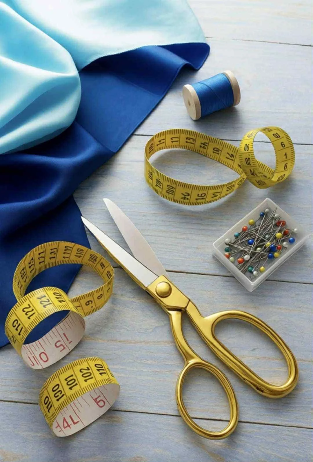 Sewing tools including a pair of gold scissors, measuring tapes, colorful sewing pins, and fabric on a light wooden surface.