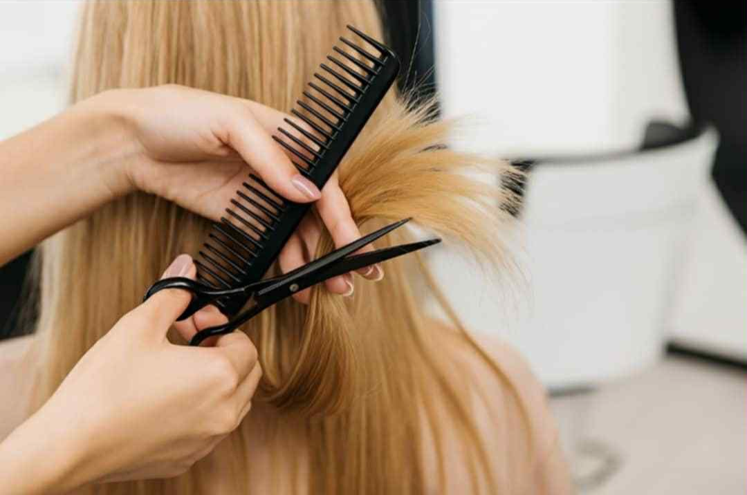 Close-up of a person cutting a client's long, blonde hair and holding a black comb and scissors in a salon setting.