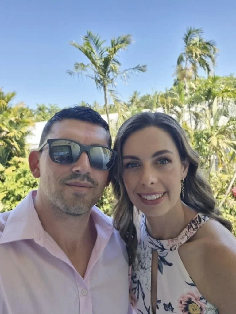 A man and woman taking a selfie outdoors with palm trees and blue sky in the background.