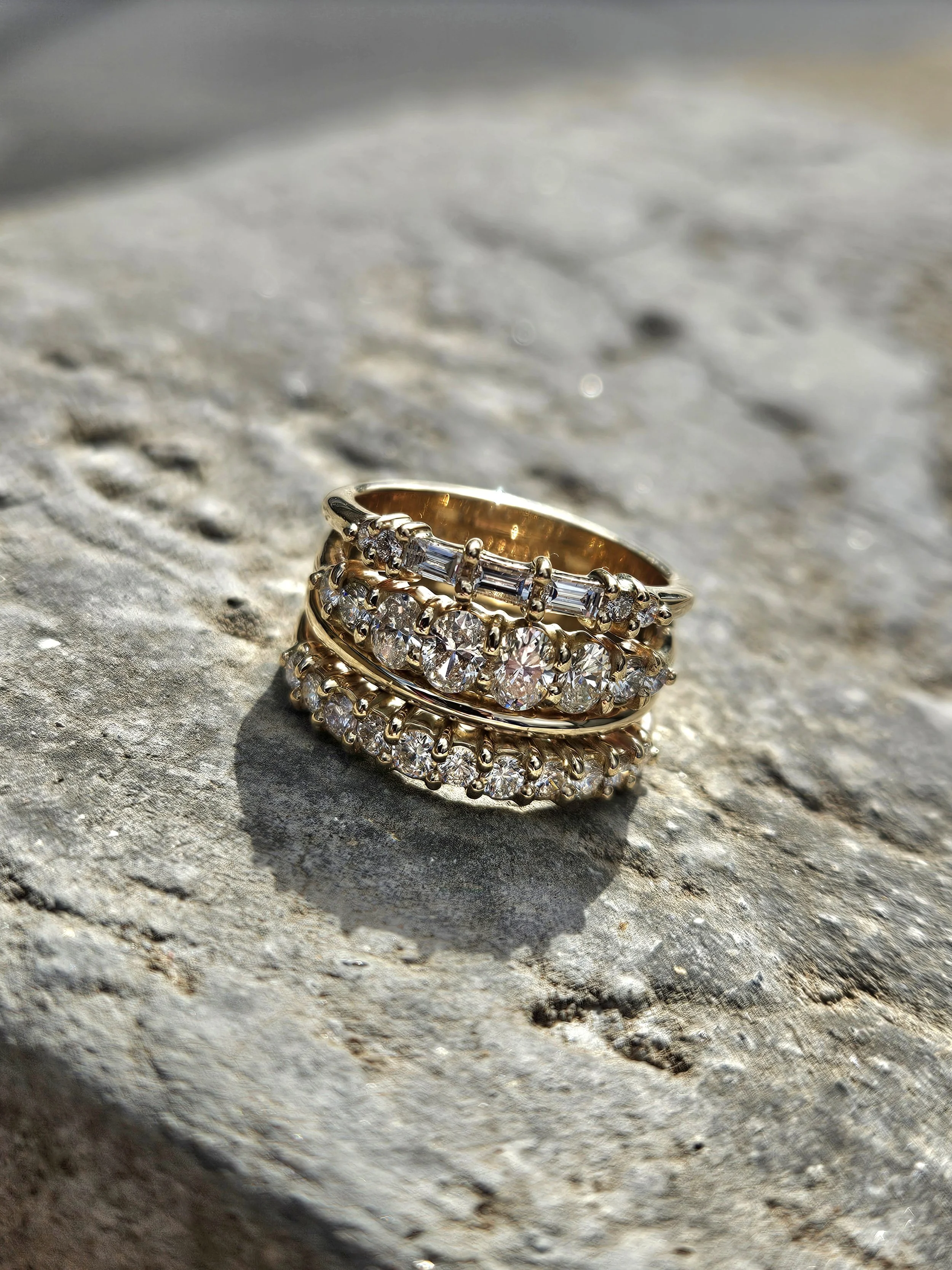 Close-up of three stacked gold rings with diamonds on a rough, textured surface, with sunlight reflecting off the rings.