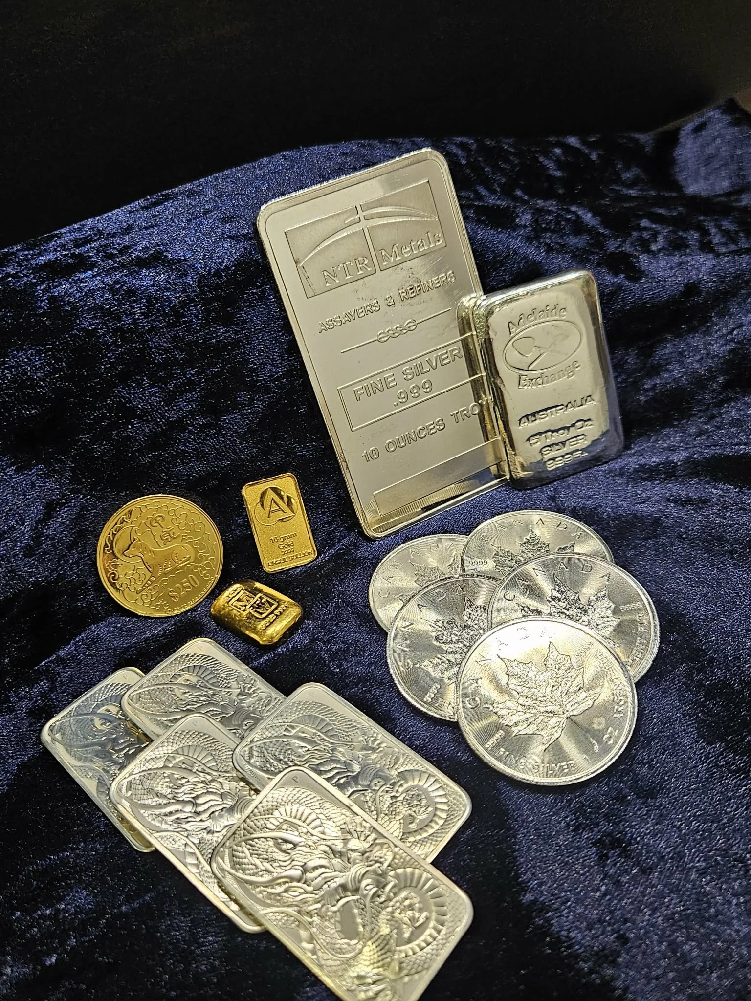 Various silver and gold coins, and silver bars on a dark fabric background.