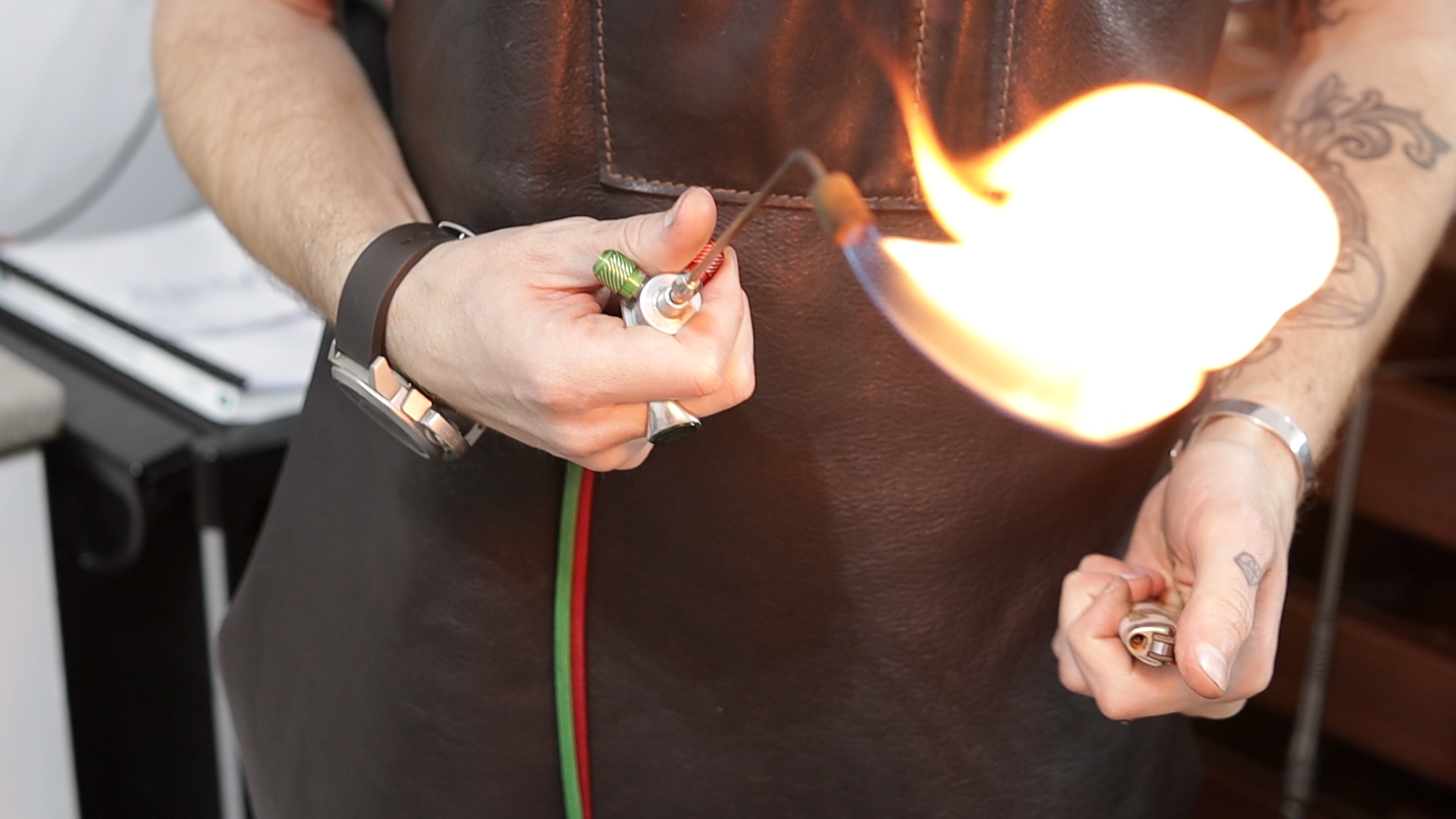 Person holding a lit blowtorch with a visible flame, wearing a brown apron and fingerless gloves.
