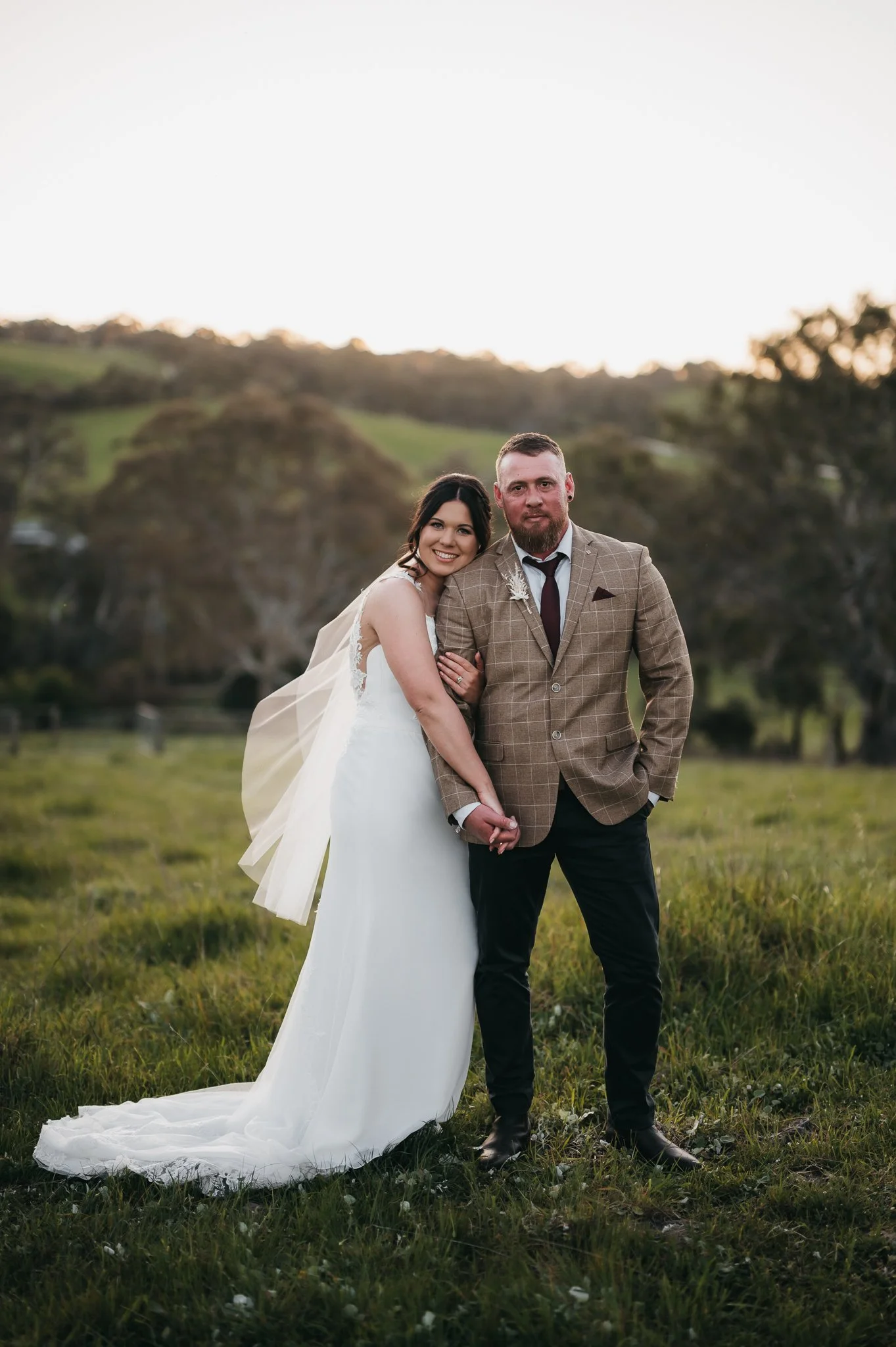 A bride and groom standing together outdoors in a grassy field with hills and trees in the background during sunset, smiling and holding hands.