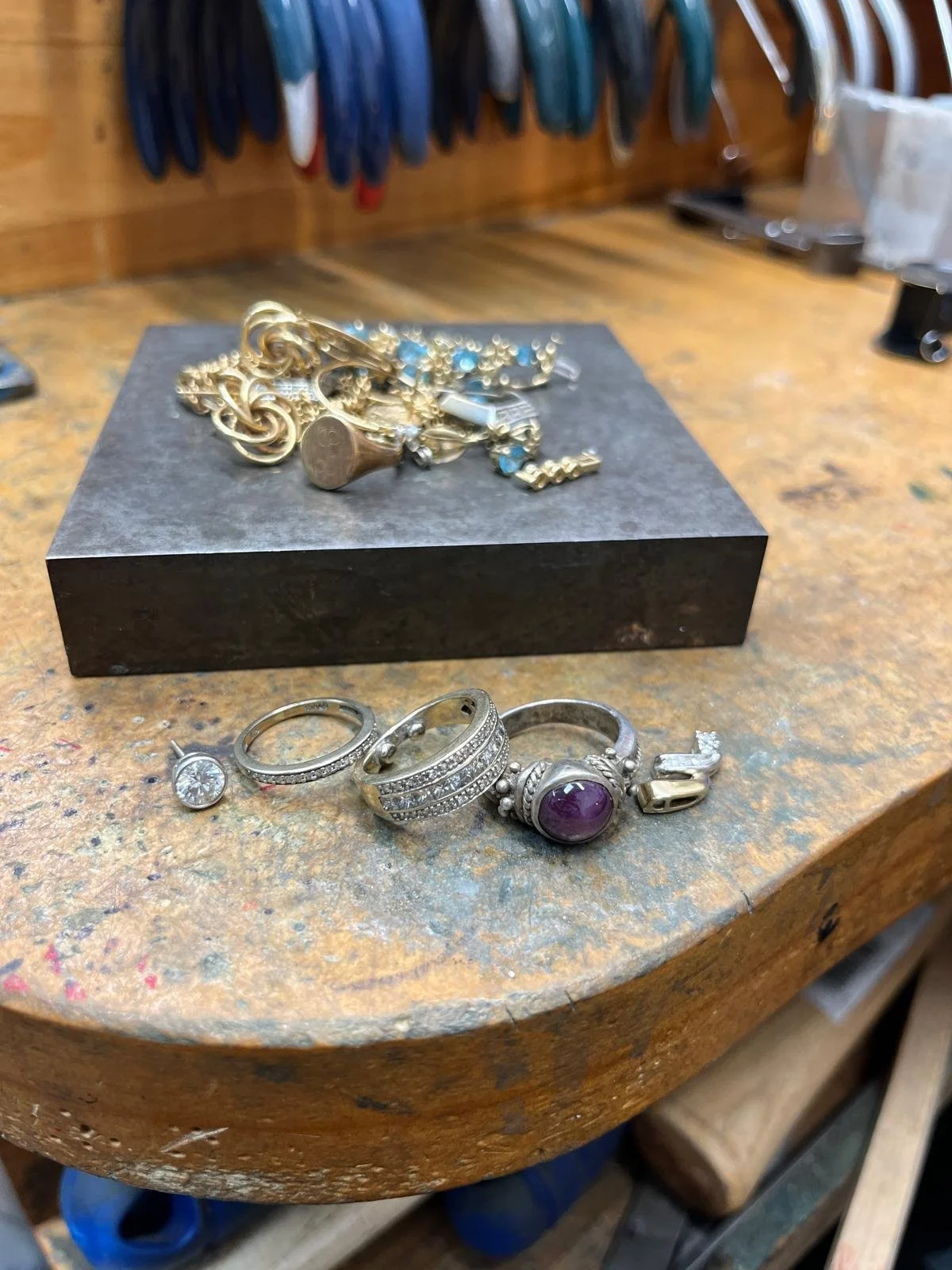 Various jewelry pieces including rings, a bracelet, and earrings on a workbench with jewelry tools in the background.