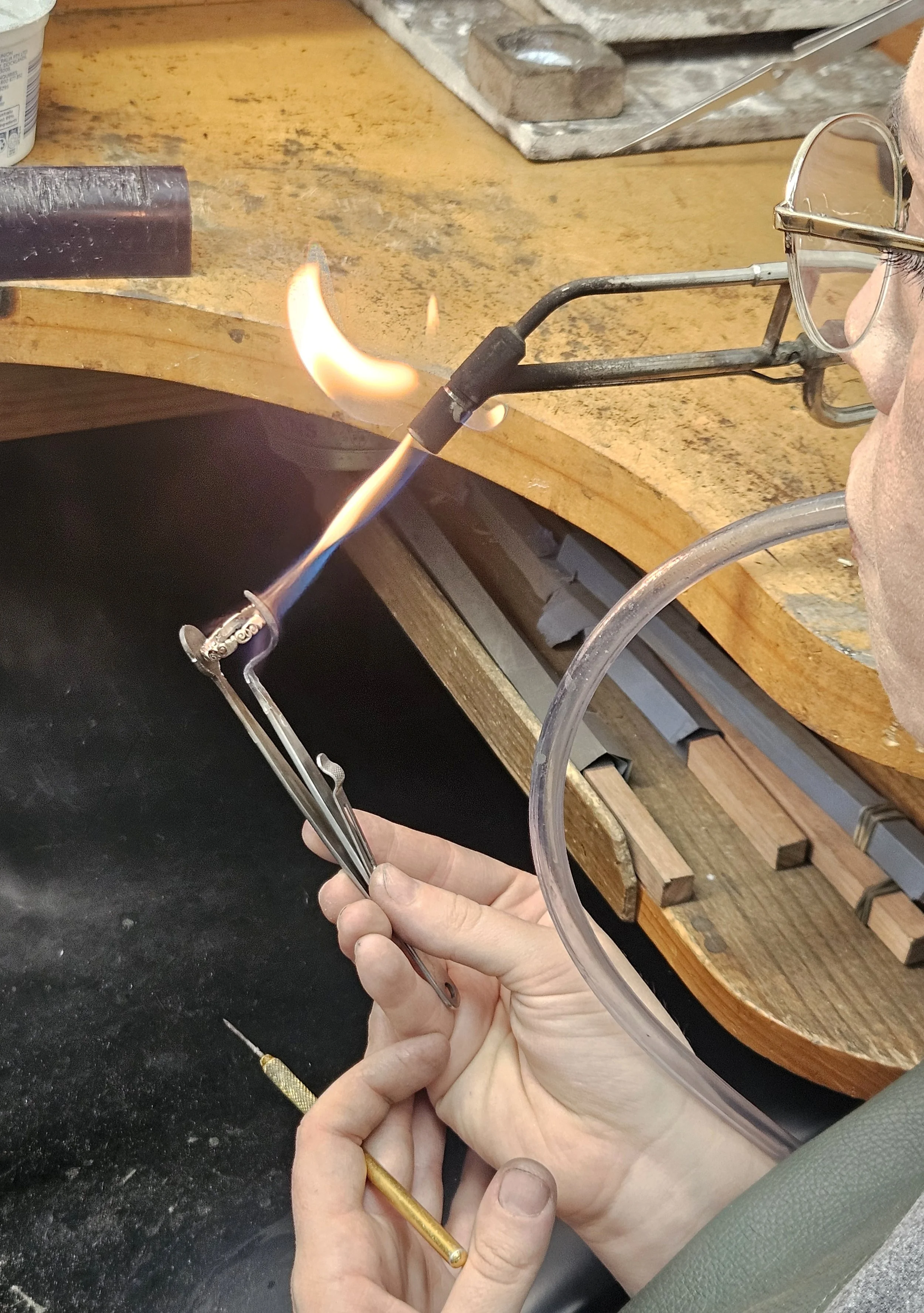 A person holding a glassblowing pipe with molten glass at the tip, working at a lampworking station with tools and equipment around.