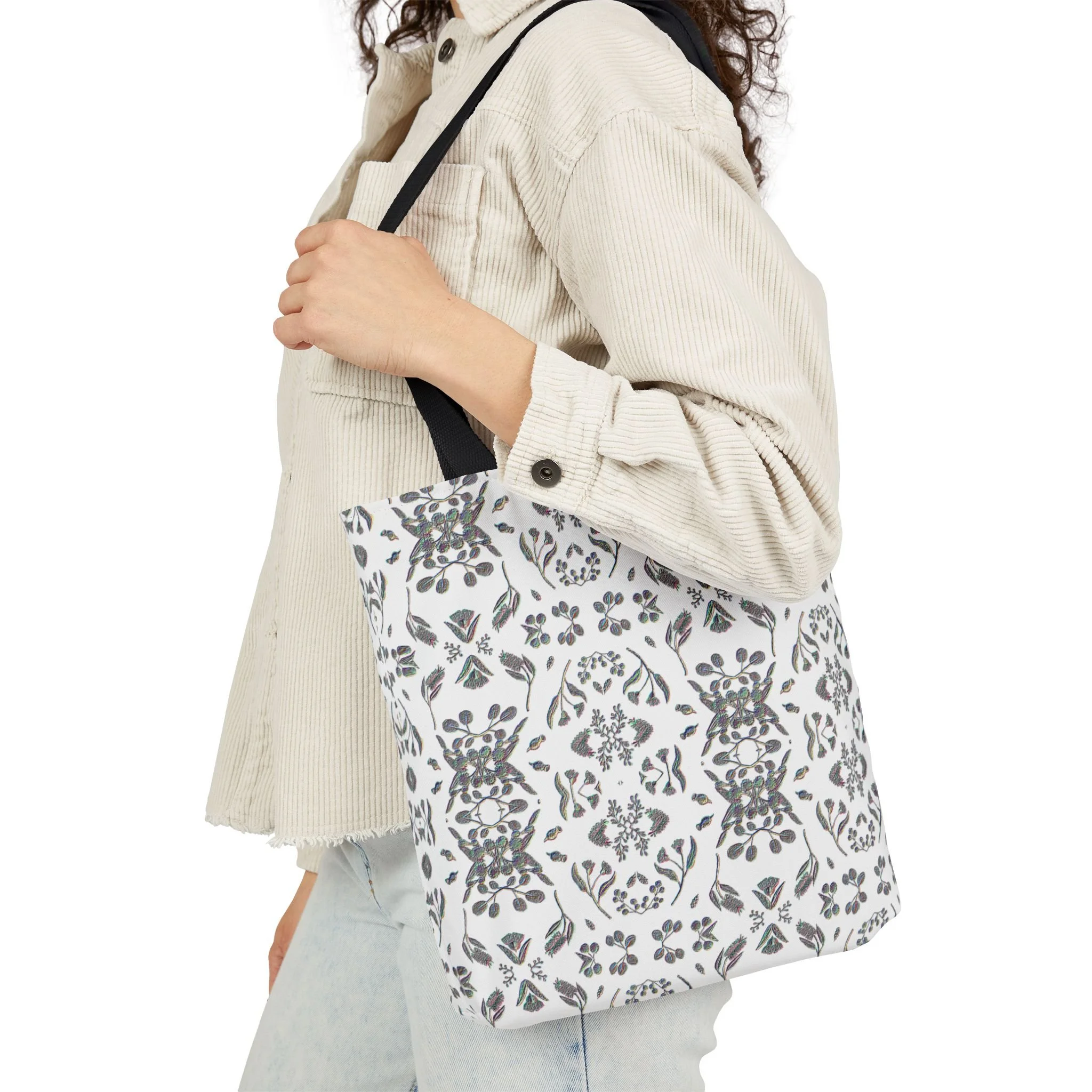 A woman carrying a white tote bag with black floral and geometric patterns, wearing a cream-colored corduroy jacket and light-colored jeans.