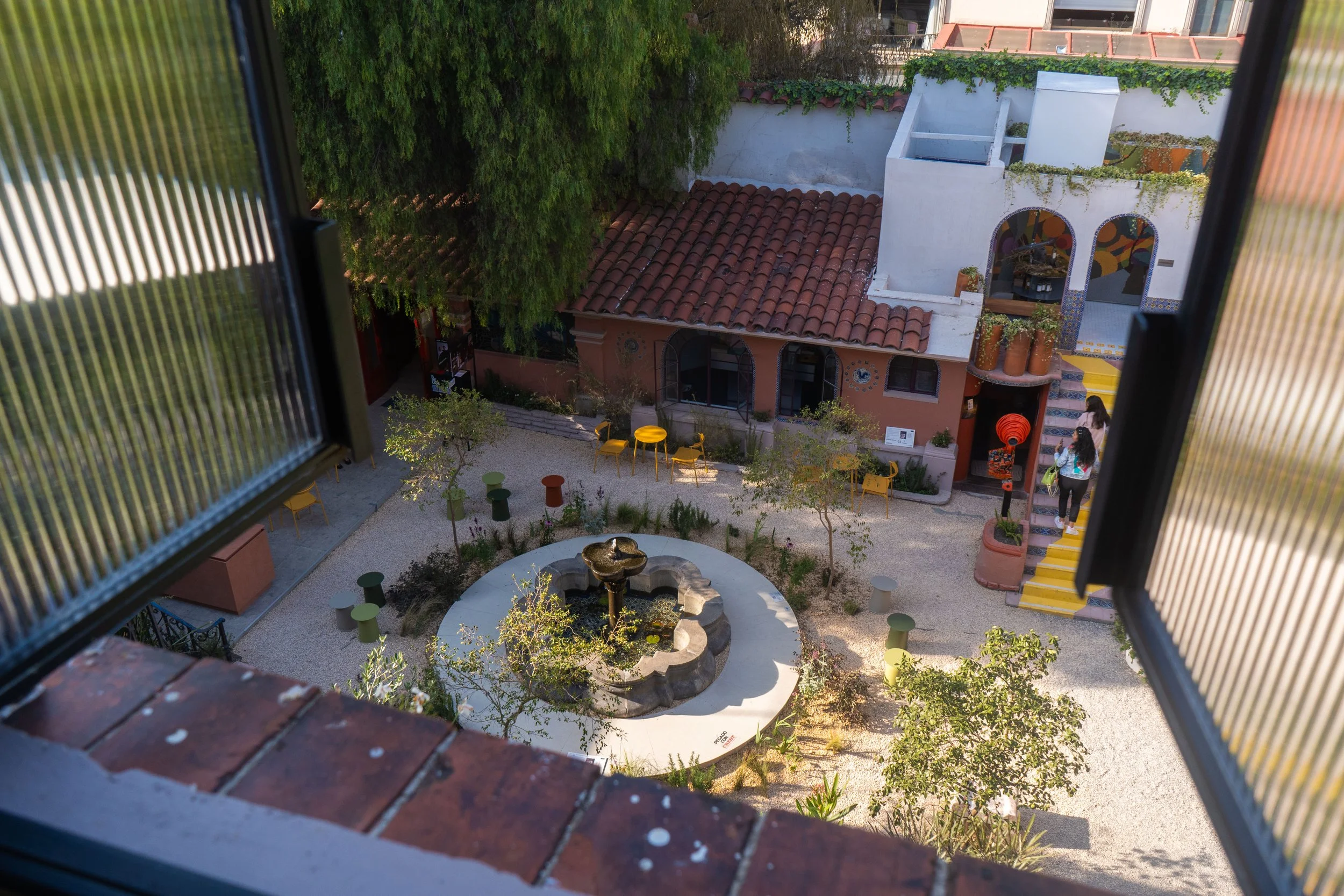 Vista de un patio interior visto desde una ventana enmarcada por contraventanas abiertas. En el centro hay una fuente decorativa, rodeada de bancos, mesas y sillas coloridas. Hay árboles y plantas alrededor del patio, y en el fondo se observa un edificio con diseño tradicional y detalles coloridos, así como dos personas entrando por unas escaleras