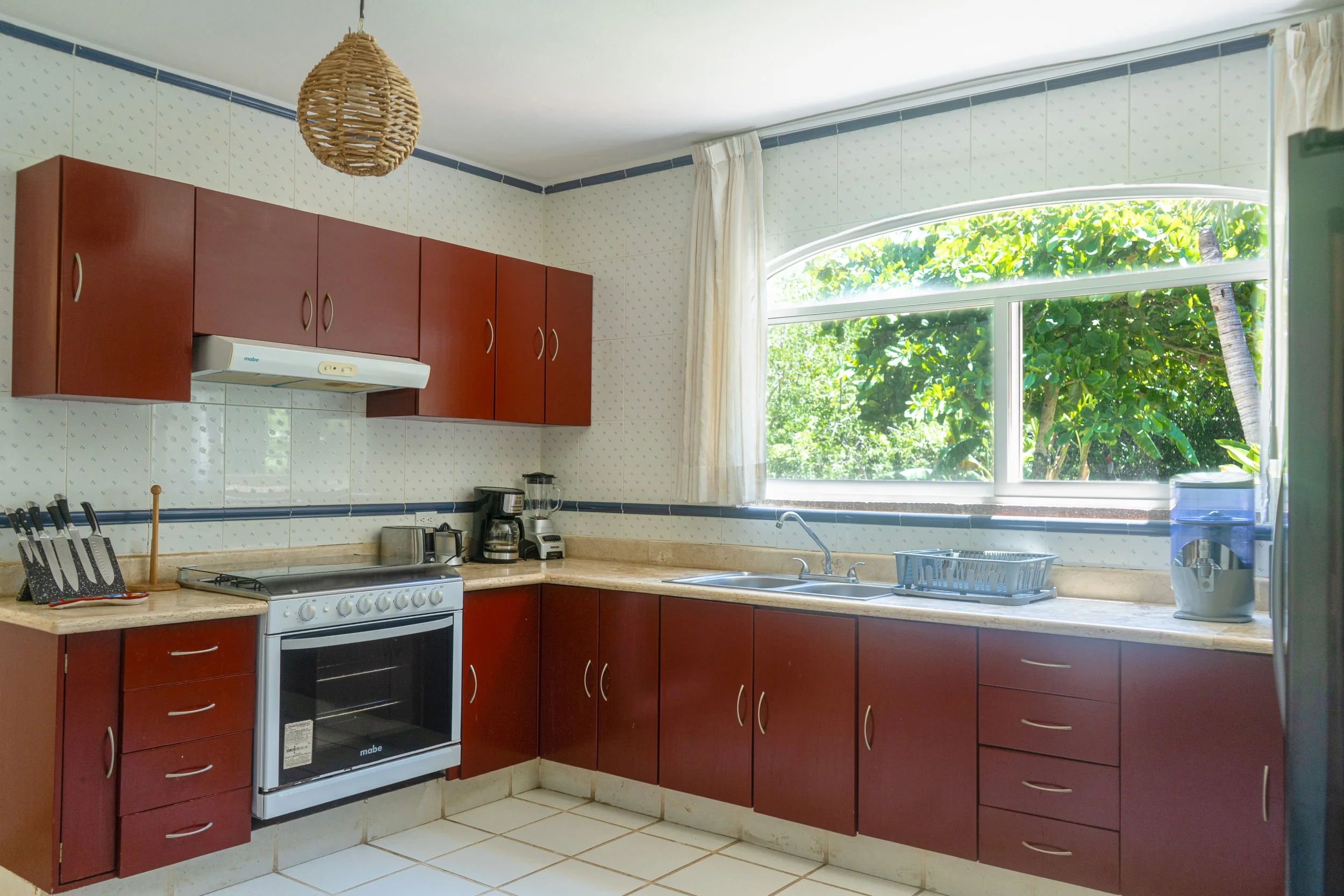 Cocina con gabinetes de madera marrón, estufa blanca, encimera de piedra, utensilios de cocina, y vista a un jardín verde a través de una gran ventana.
