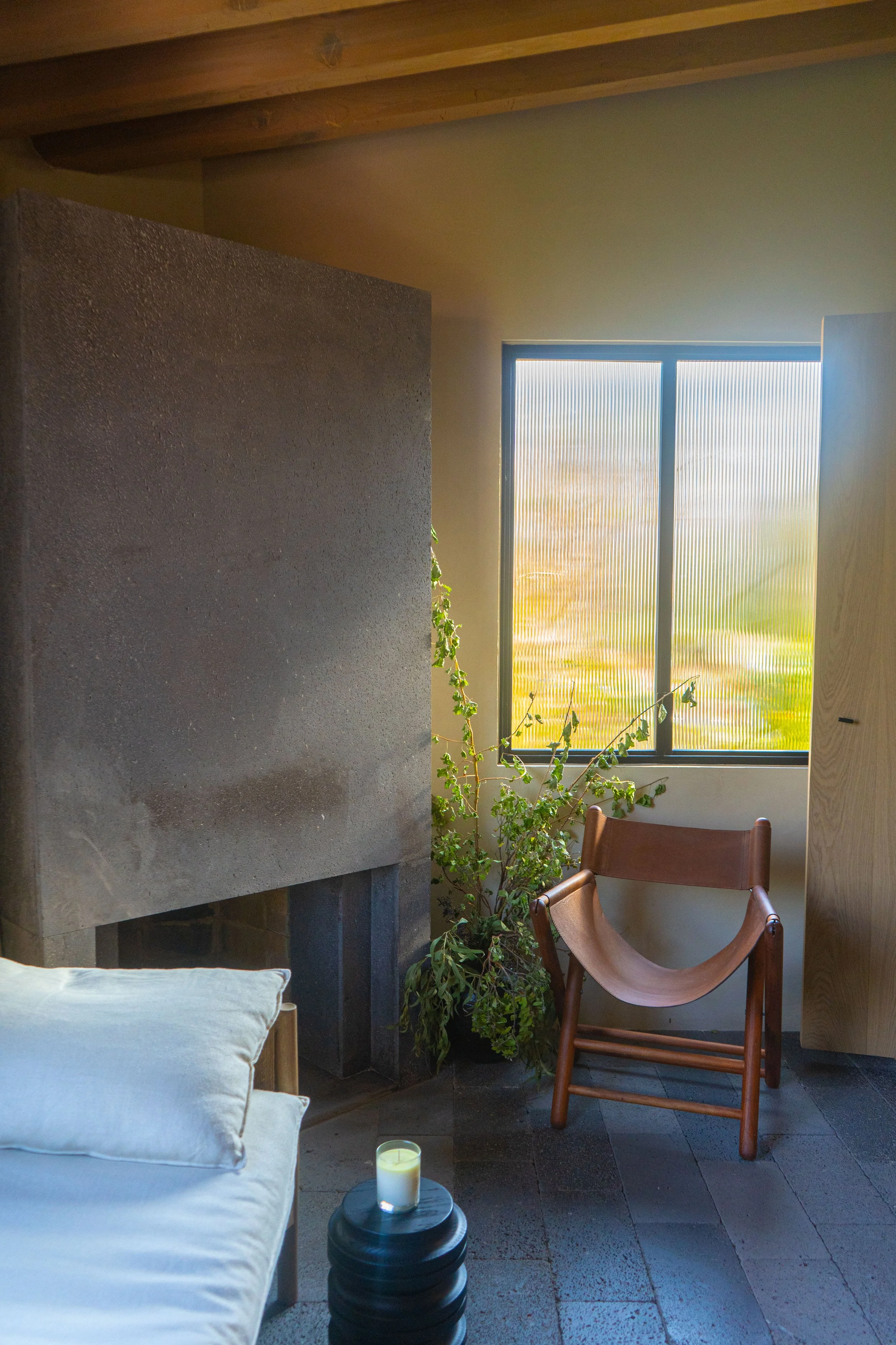 Interior de una habitación con pared de piedra, ventana con vista exterior, planta, silla de madera con cuero y cama con almohada, además de una vela en un soporte decorativo.