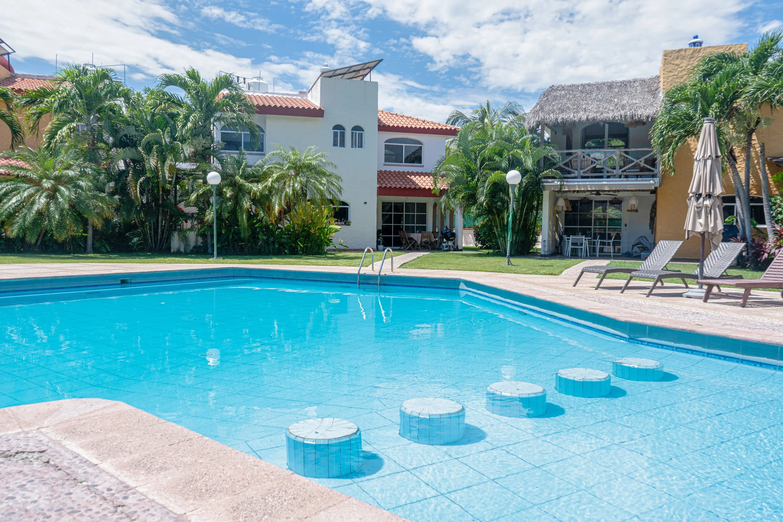 Piscina en un complejo residencial con casas coloridas, palmeras, sillones y sombrillas, bajo un cielo despejado.