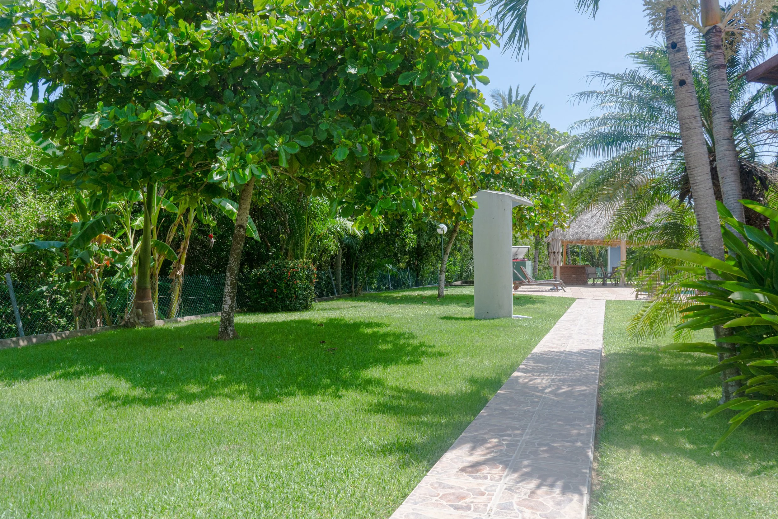 Jardín soleado con césped verde, árboles frondosos, palmeras, camino de piedra y área de descanso con camastro y sombra al fondo.