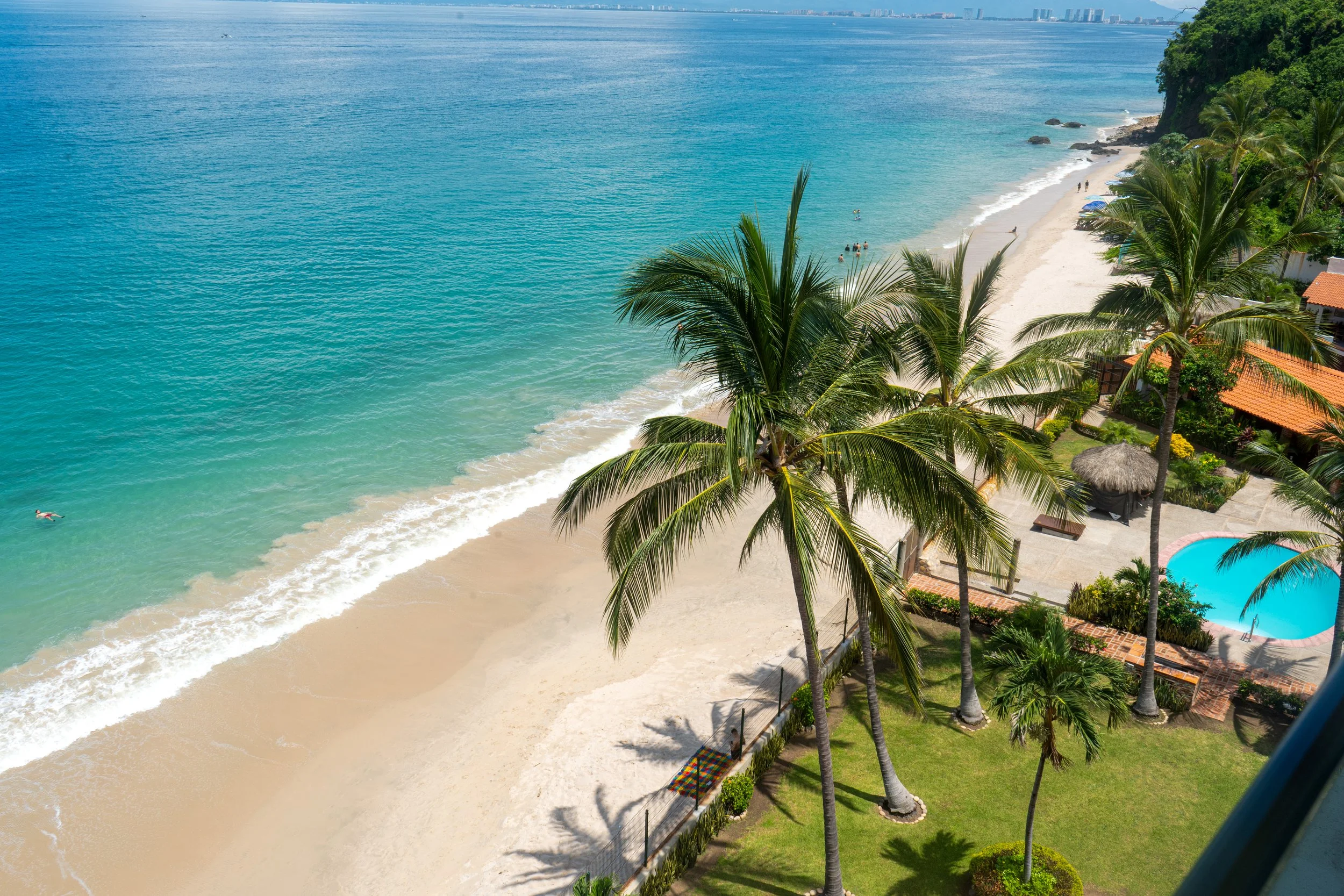 Vista aérea de una playa en el mar Caribe con palmeras, arena blanca, una piscina y casas rodeadas de vegetación.