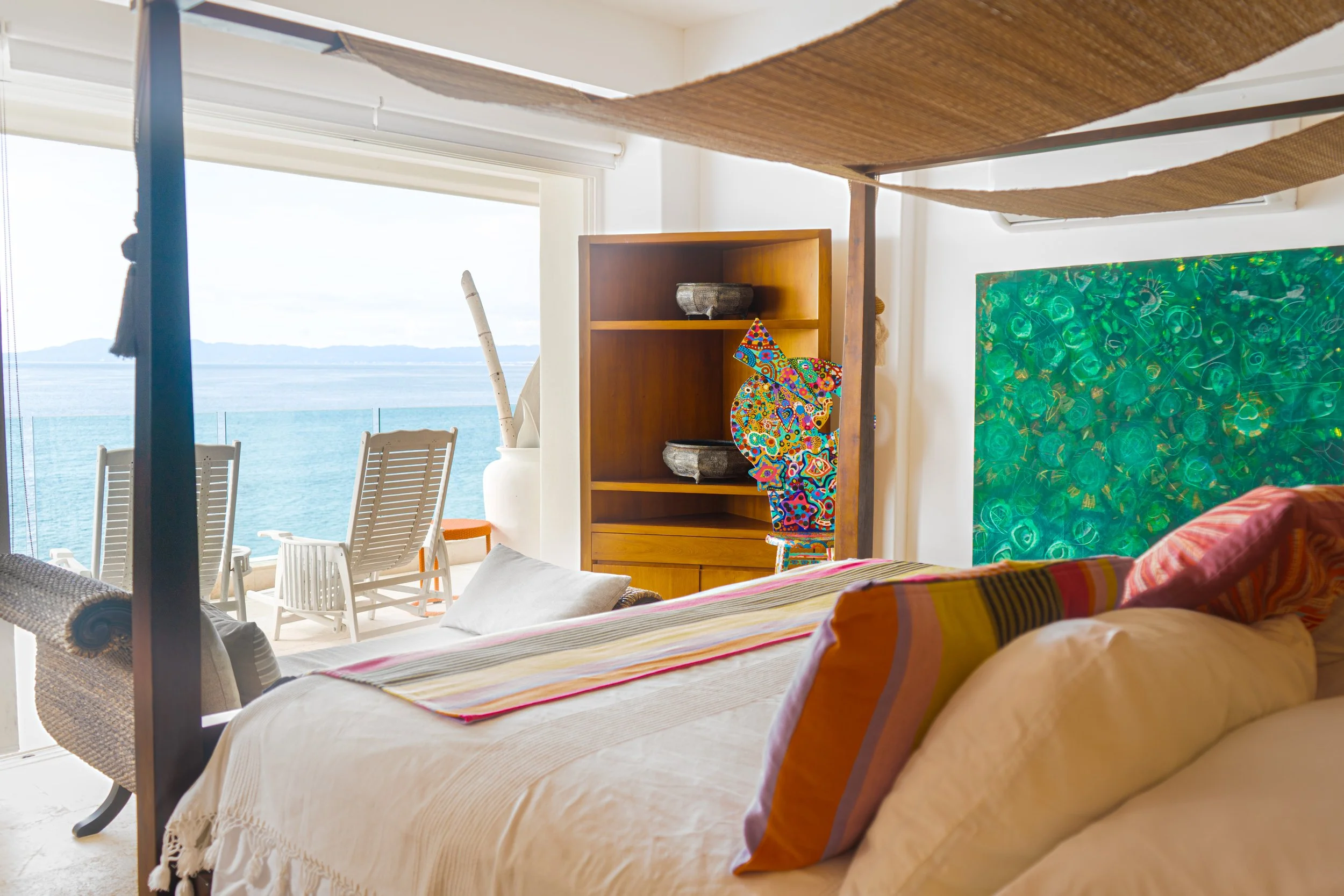 Habitación con vista al mar, con cama, cojines, una estantería de madera con decoraciones y una ventana grande con vista a la playa con sillas.