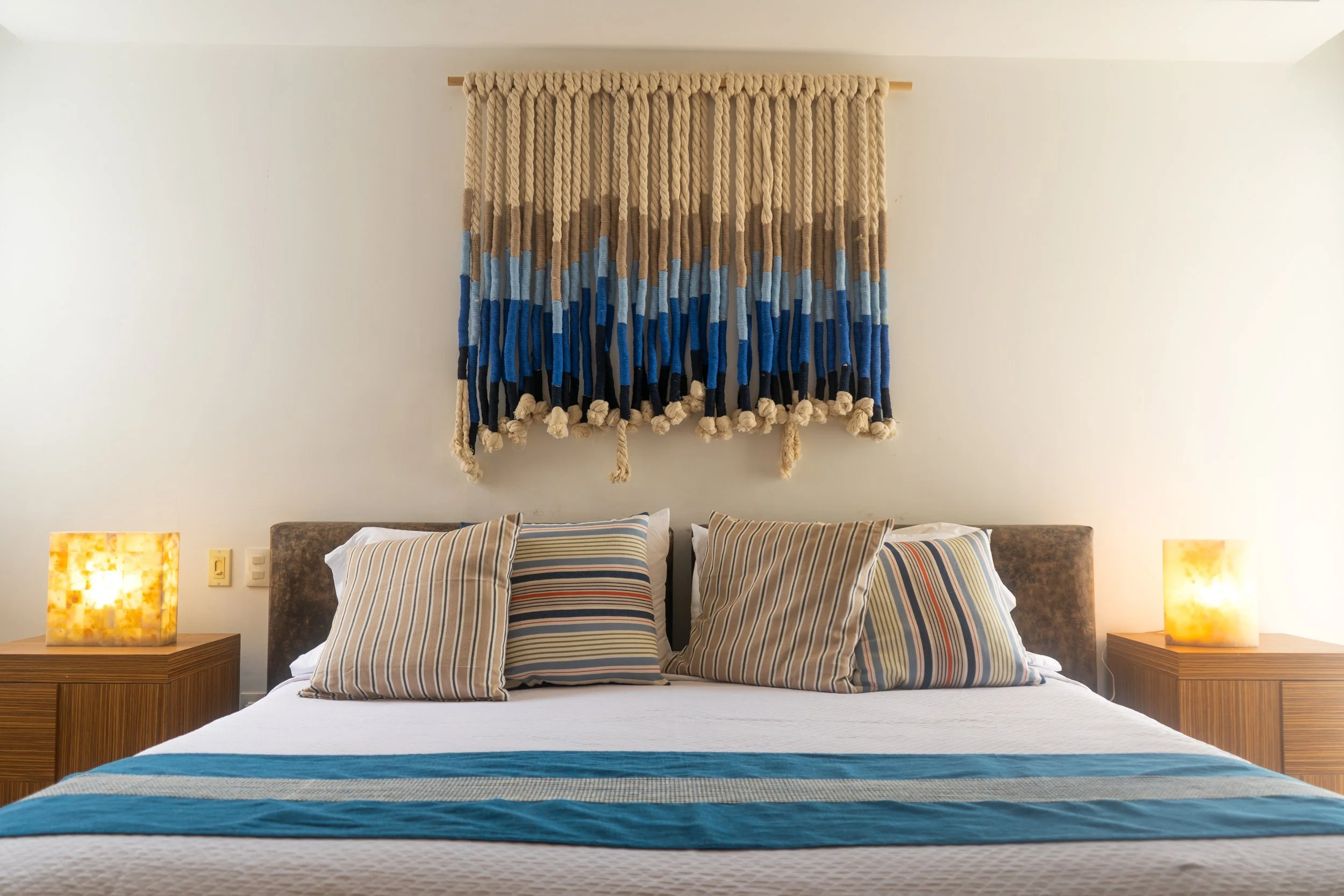 Cama con almohadas a rayas en un dormitorio, con lámparas de mesa en ambas esquinas y tapiz de macramé en la pared del fondo.