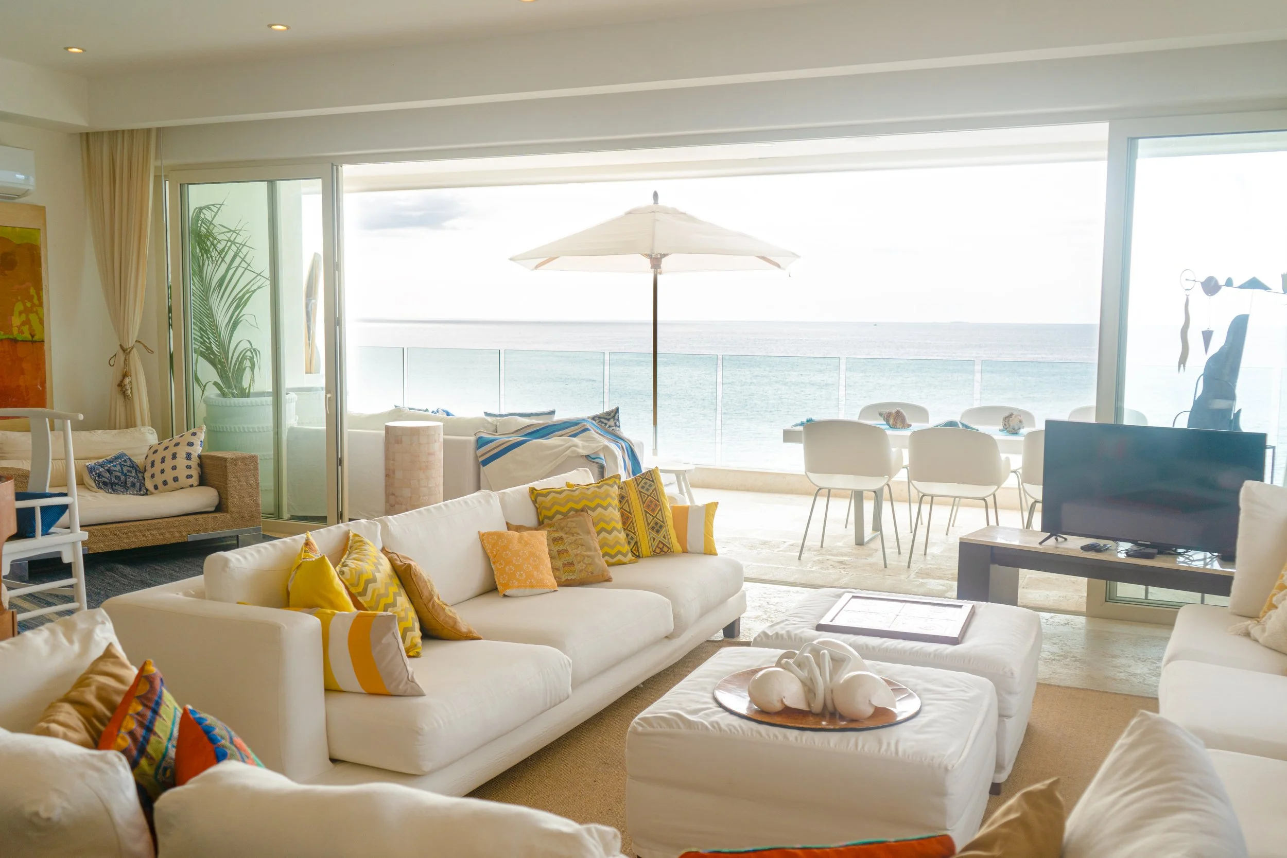 Sala de estar con vista al mar, muebles blancos con cojines coloridos y una terraza con sombrilla y comedor junto a la barandilla.