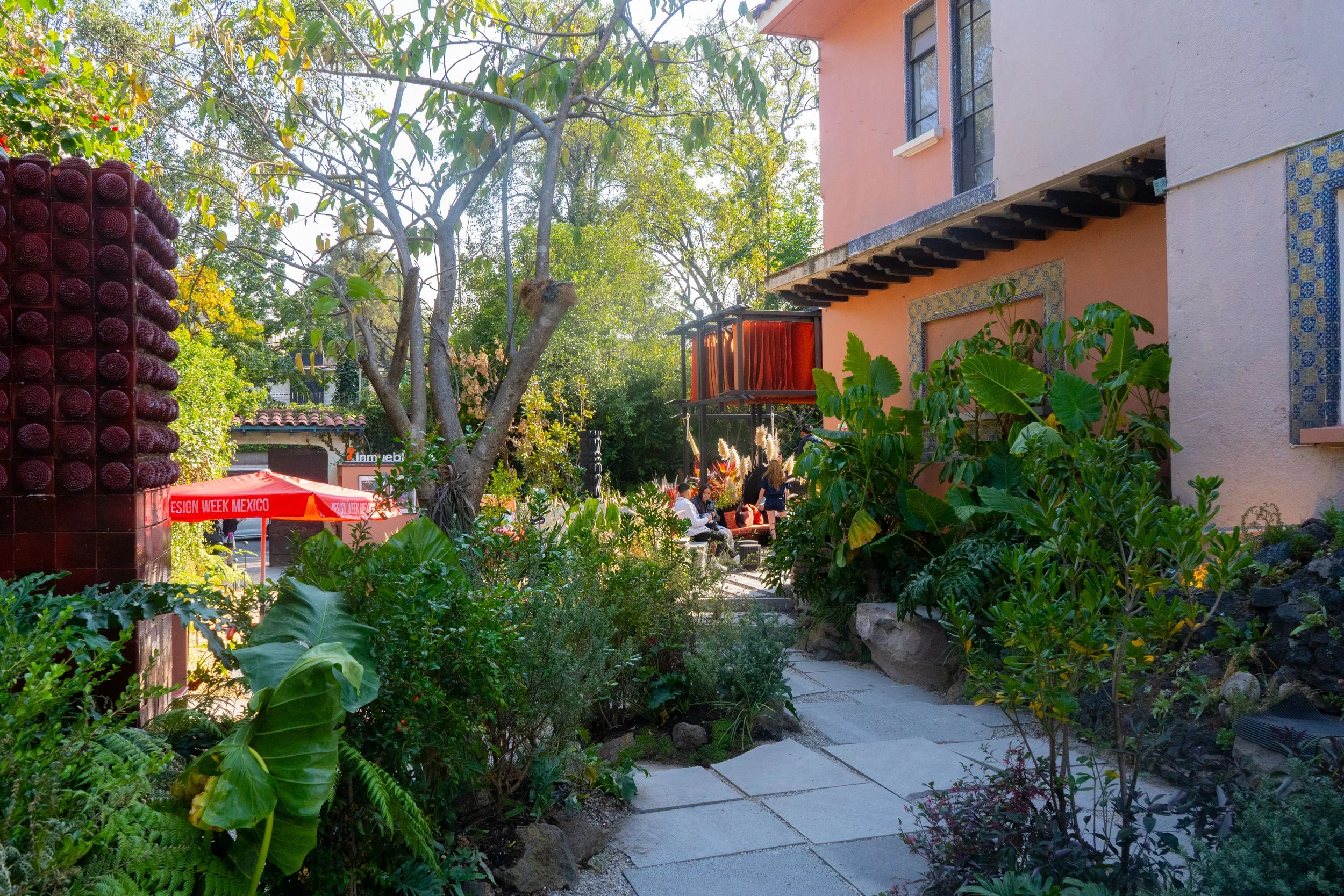 Zona de jardín con plantas verdes, árboles y un camino de piedra frente a una casa de color rosa con ventanas de barro y una terraza con personas sentadas. En el fondo hay un cartel que dice 'ESIGN WEEK MEXICO' y una estructura decorativa con botellas.